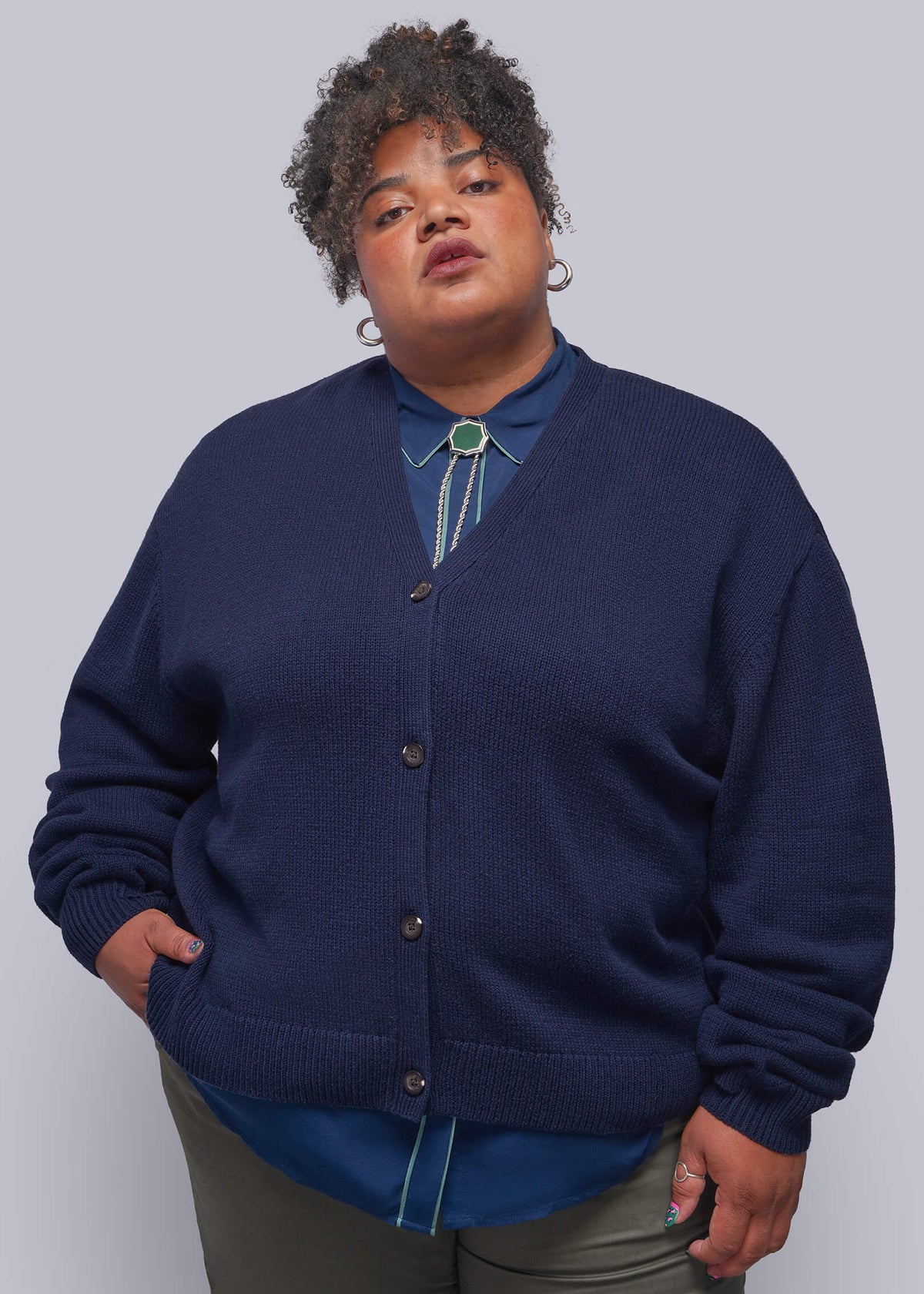 1X=A plus-size model wearing a navy blue knit cardigan over a blue collared shirt with a turquoise bolo tie, paired with olive green pants, against a solid gray studio background.