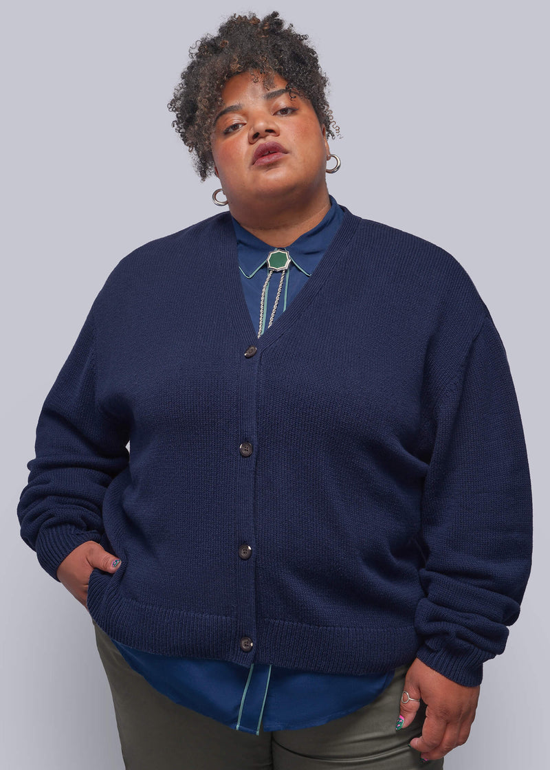 1X=A plus-size model wearing a navy blue knit cardigan over a blue collared shirt with a turquoise bolo tie, paired with olive green pants, against a solid gray studio background.