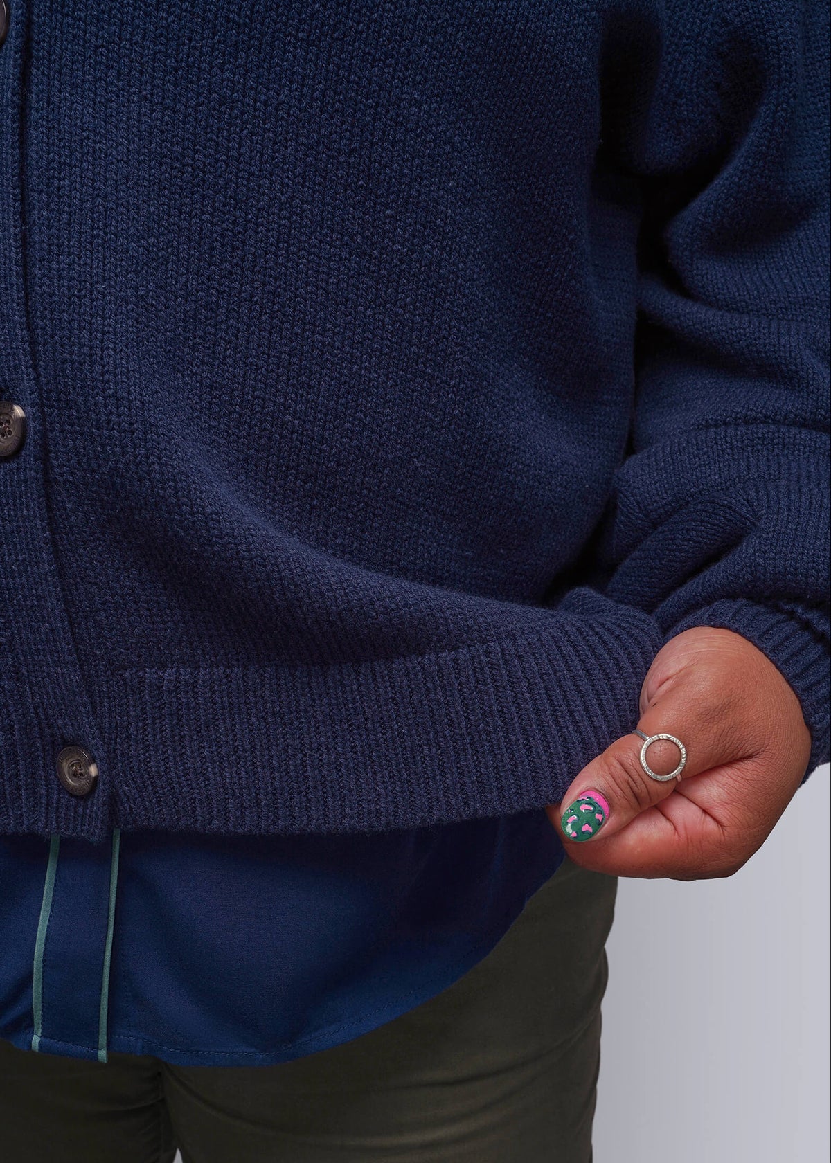 1X=Close-up of a person wearing a navy blue knit cardigan over a blue top and olive green pants, with one hand holding the hem of the cardigan.