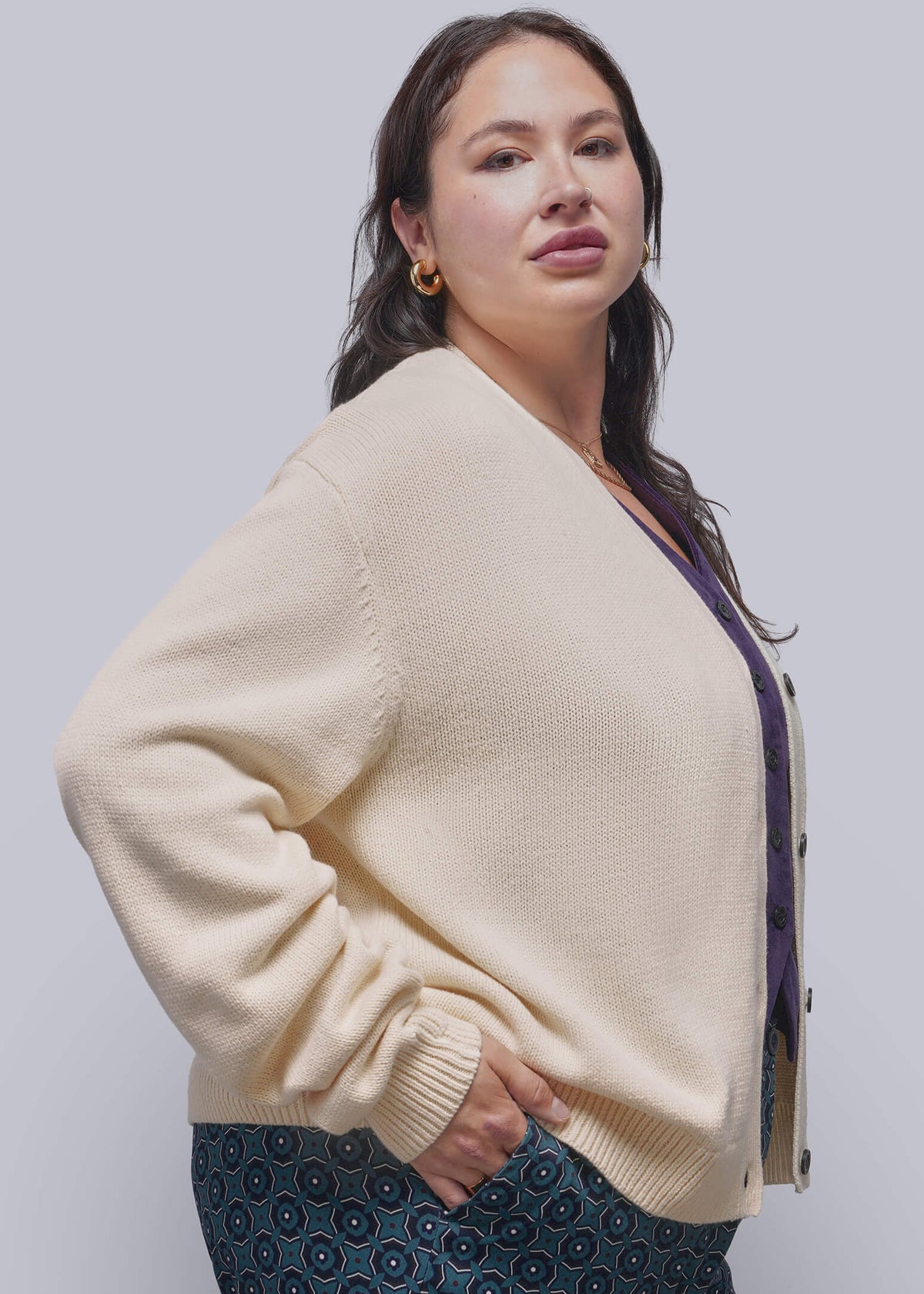 S=A woman modeling a cream-colored knit cardigan sweater with a purple inner layer and dark patterned pants, photographed against a solid light gray studio background.