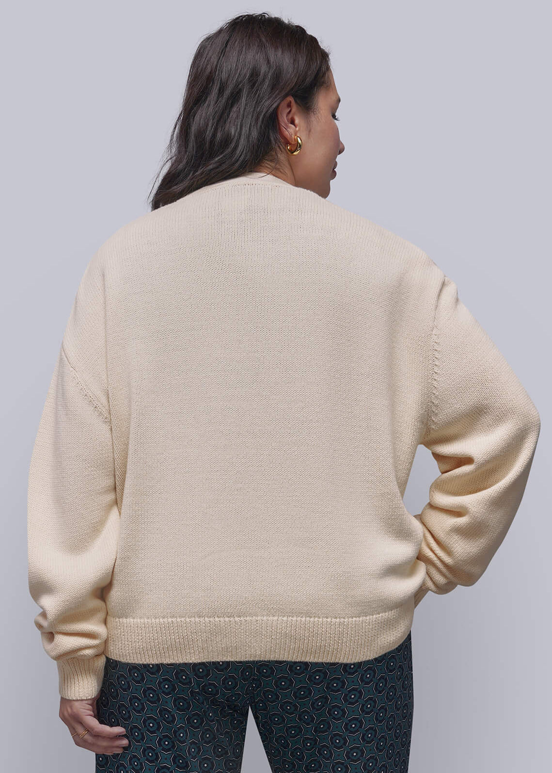 1X=A model shown from the back wearing a cream-colored knit sweater and dark patterned trousers, standing against a light gray studio background.
