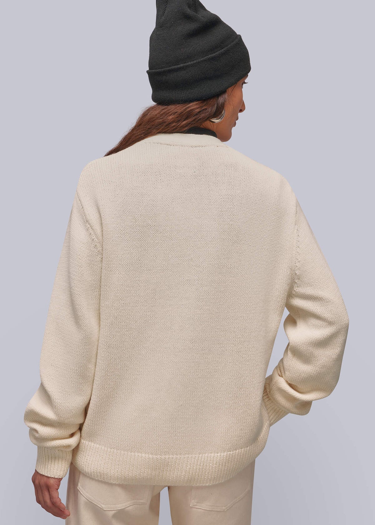 S=A person shown from the back wearing a cream knit sweater and matching cream pants, with a black beanie, against a light gray studio background.
