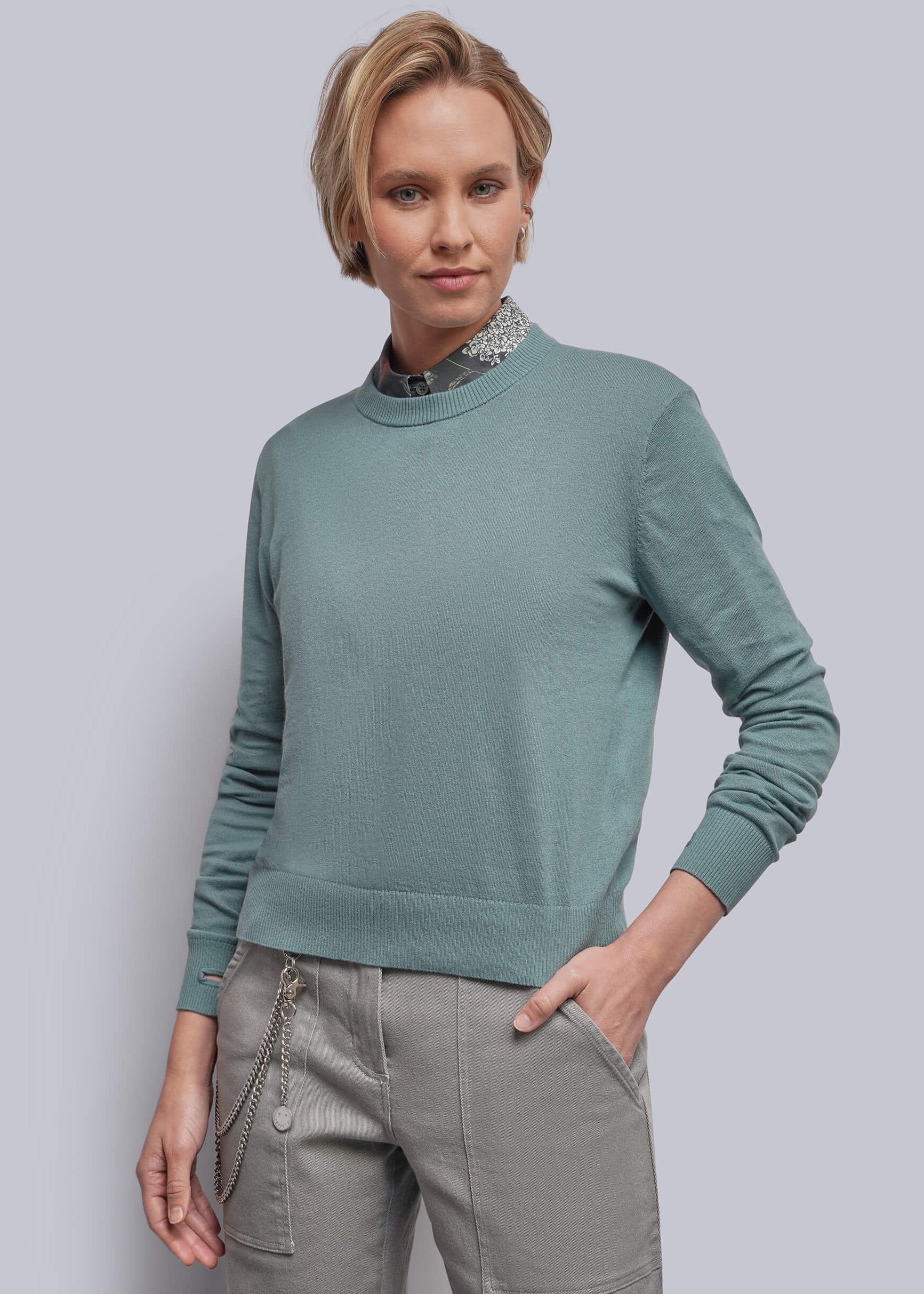 S=A woman modeling a teal long-sleeve crewneck sweater paired with gray pants, standing against a solid light gray background.