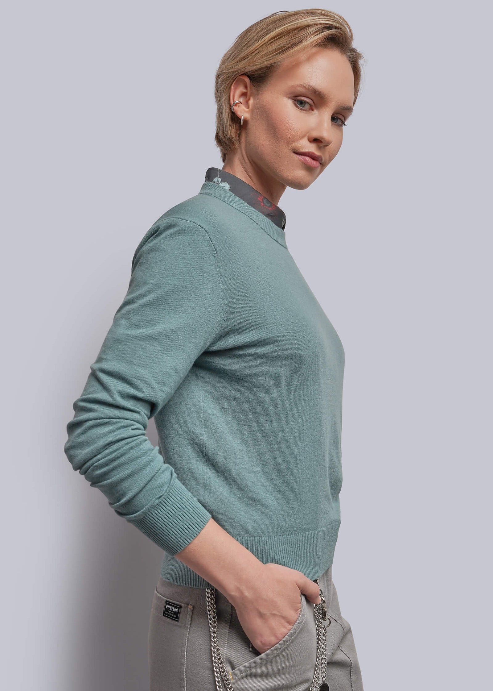 S=A woman wearing a teal long-sleeve crewneck sweater and light gray trousers, standing against a solid light gray studio background.