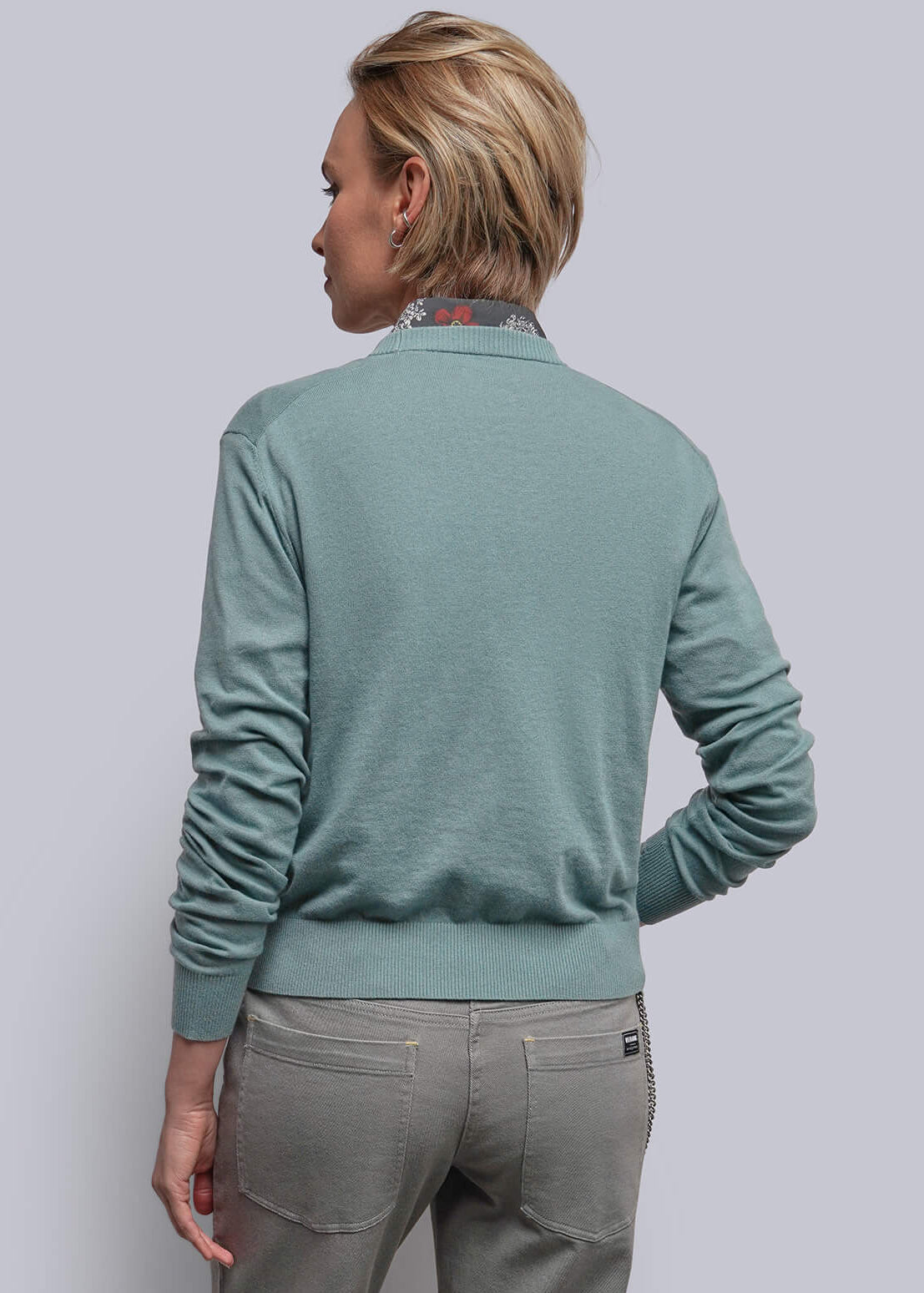 S=A person viewed from behind wearing a teal long-sleeve knit sweater and light gray trousers, standing against a plain light gray wall.