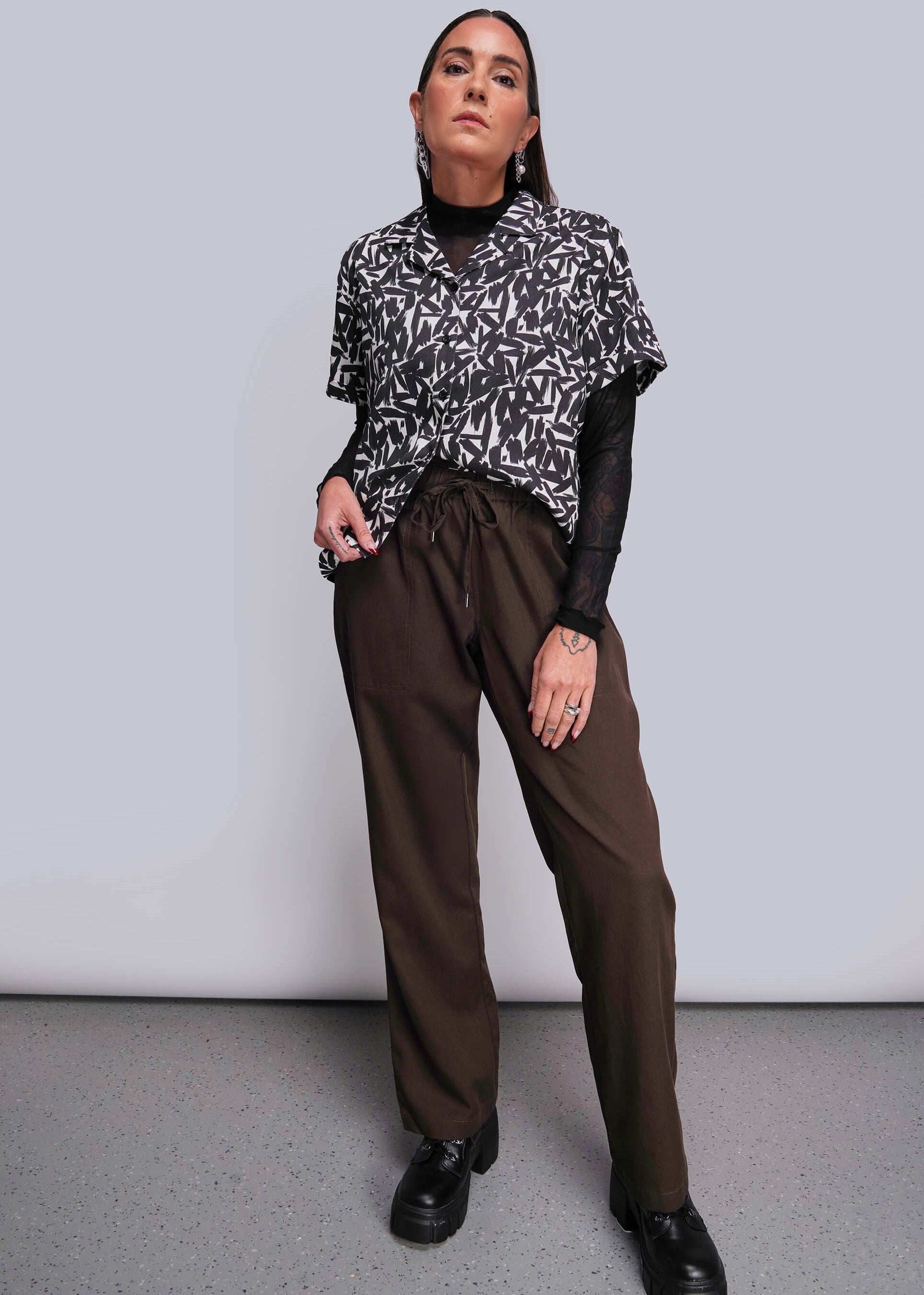 Person wearing a patterned black and white top with brown pants against a plain background