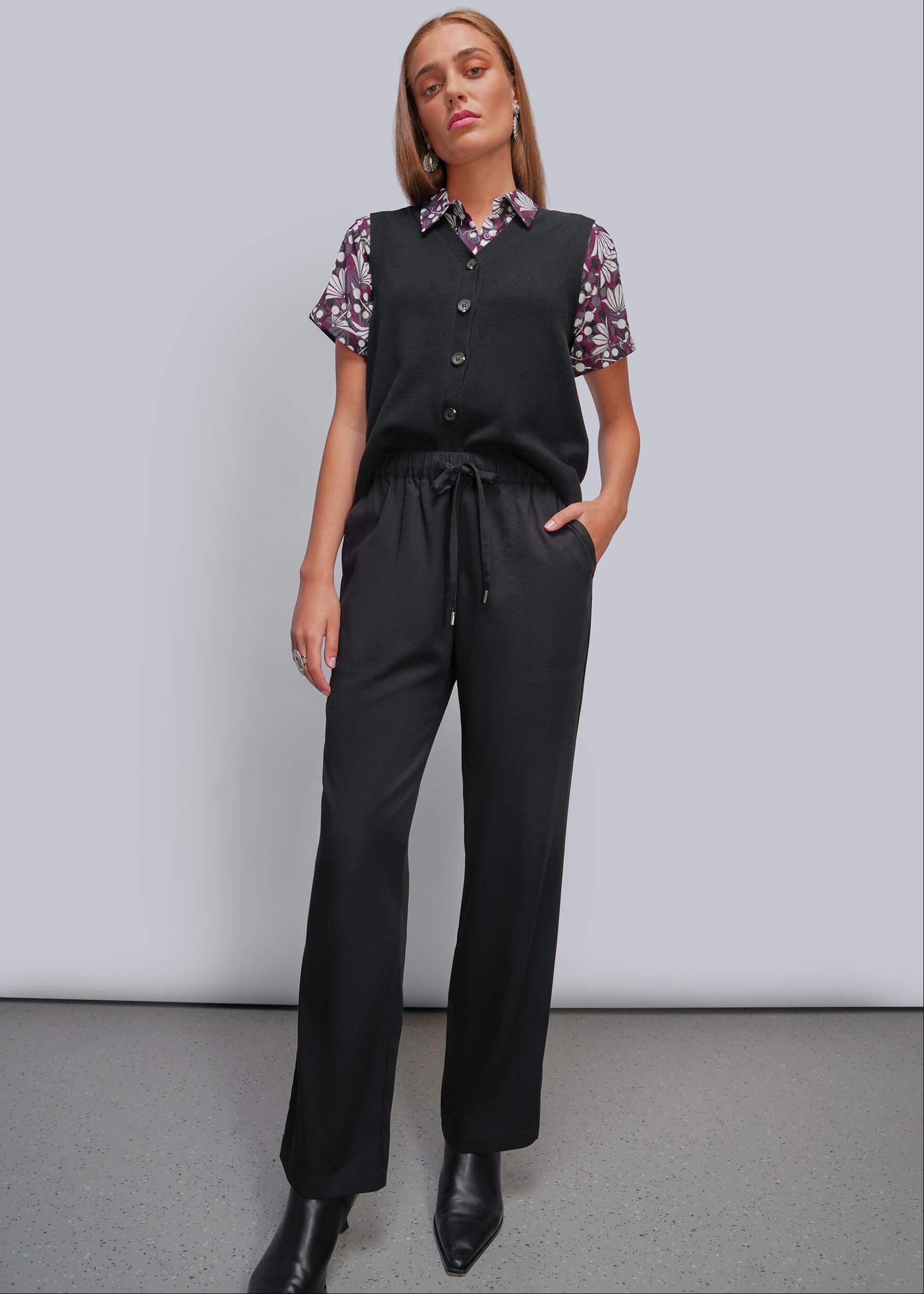 A woman modeling a black sleeveless vest and matching black drawstring trousers over a short-sleeve floral collared shirt, standing against a light gray studio background.