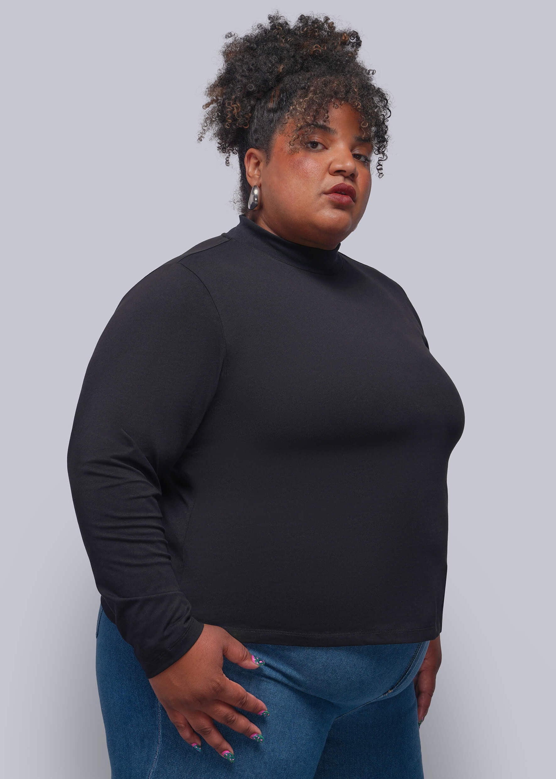 1X=A plus-size model wearing a black long-sleeve mock neck top and blue jeans, standing against a solid gray studio background.