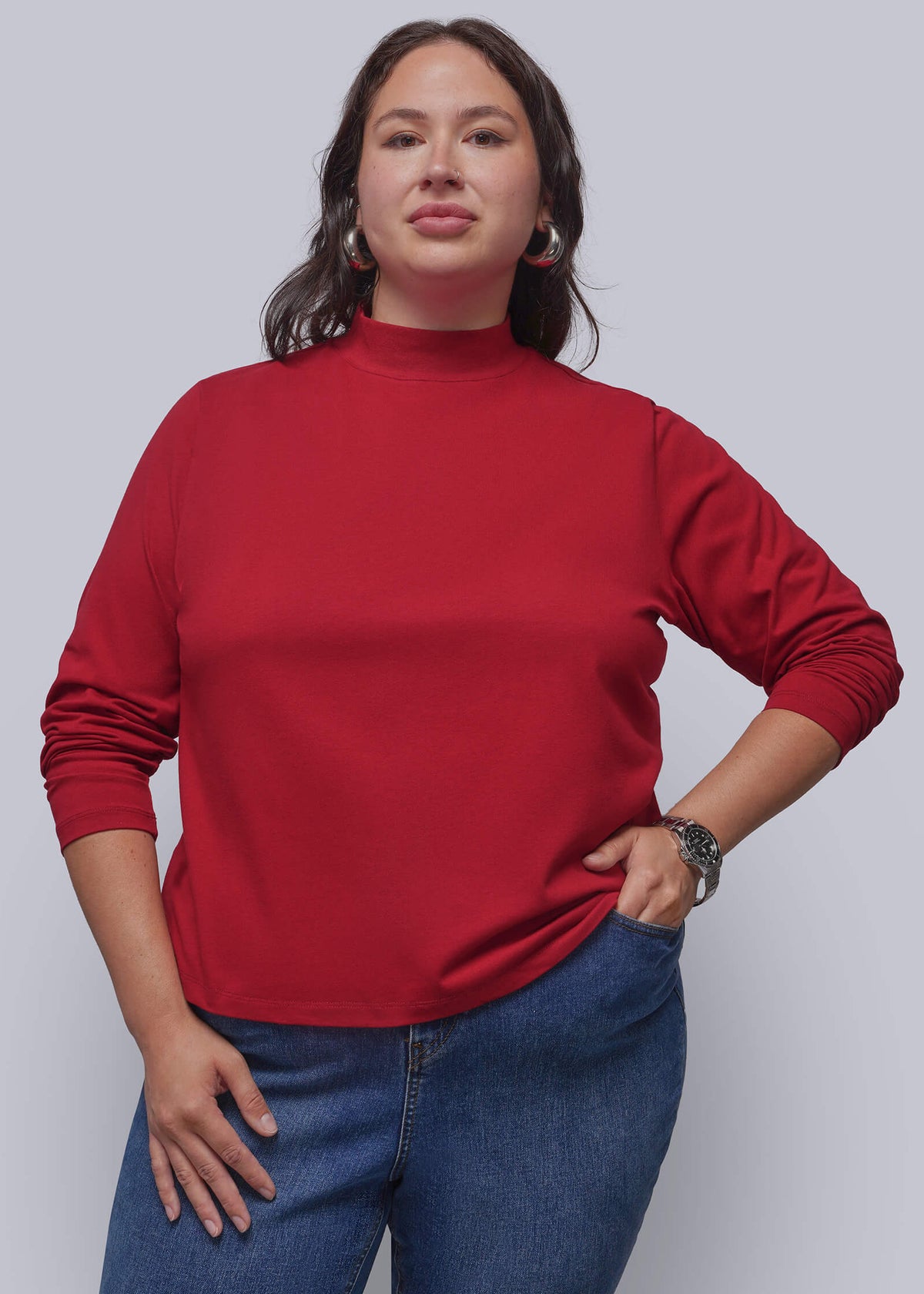 1X=A woman modeling a red mock neck long-sleeve top with blue jeans, standing against a solid gray studio background.