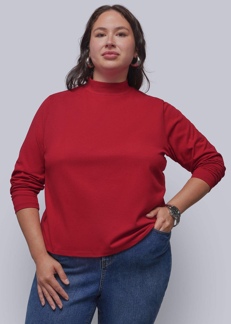 1X=A woman modeling a red mock neck long-sleeve top with blue jeans, standing against a solid gray studio background.