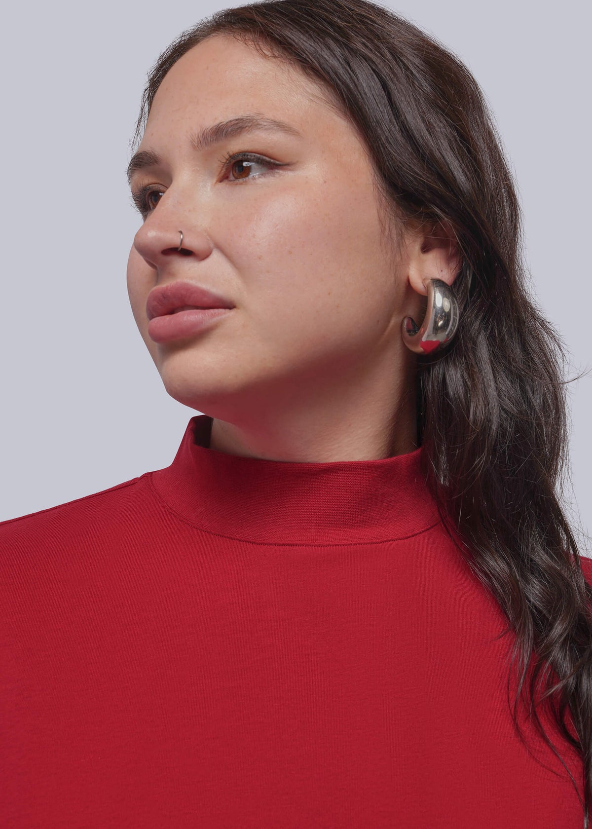 1X=A close-up of a woman wearing a bright red mock neck top and a large silver-toned geometric hoop earring, shown against a light gray studio background.