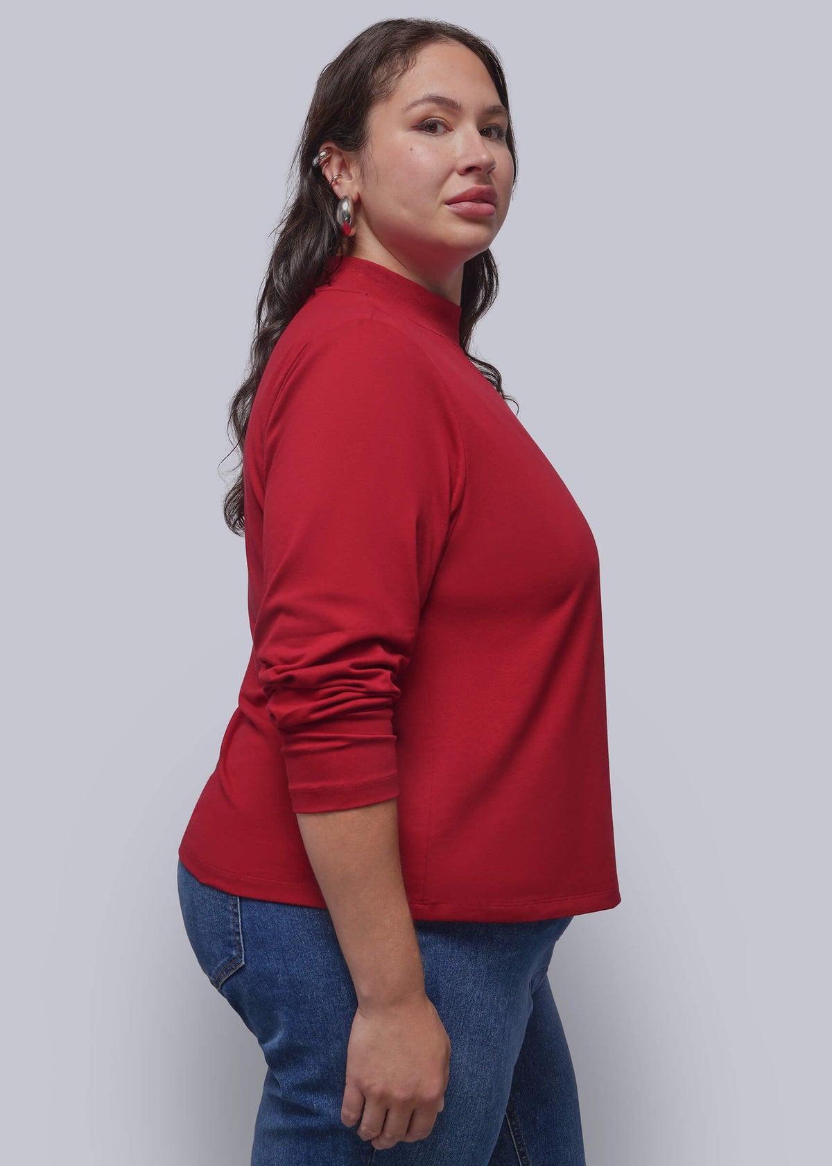 1X=A woman wearing a red mock neck long-sleeve top and blue jeans, shown in a side profile against a solid gray studio background.