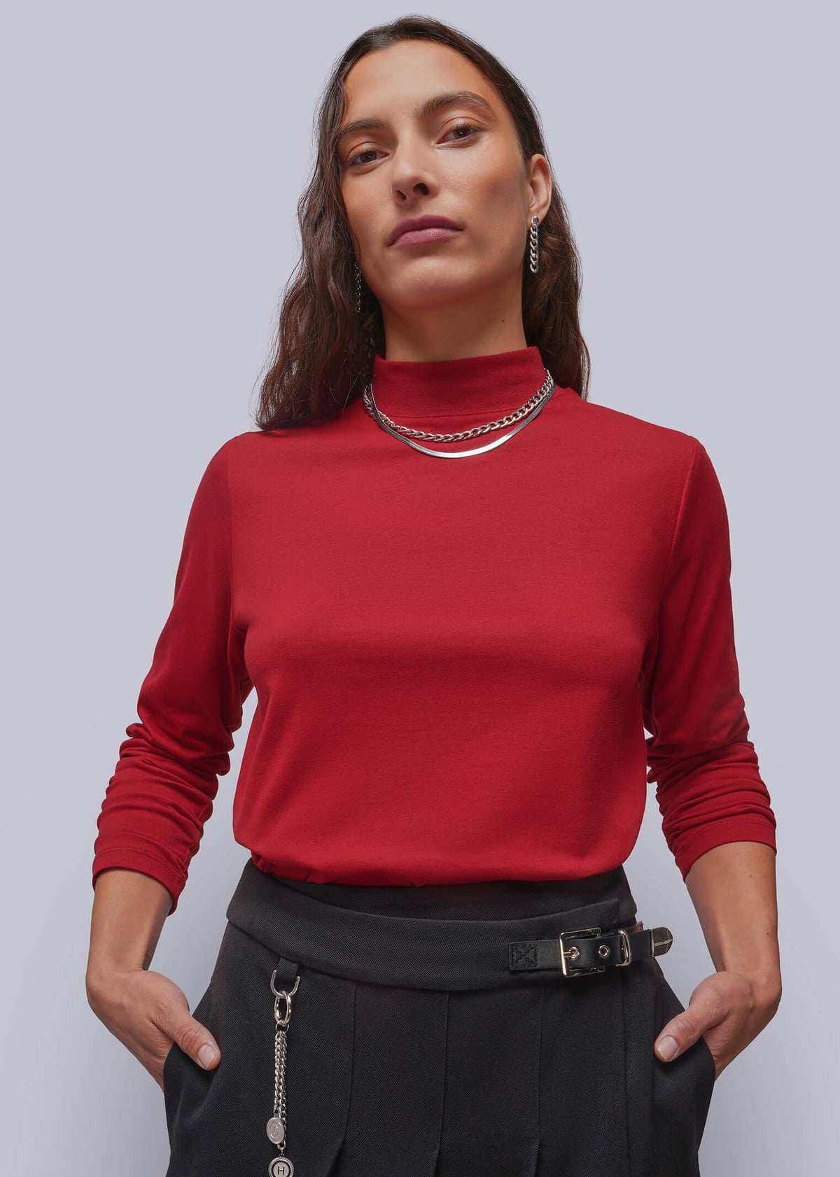S=A woman modeling a red mock-neck long-sleeve top paired with black pleated skirt and silver jewelry, set against a solid light gray studio background.
