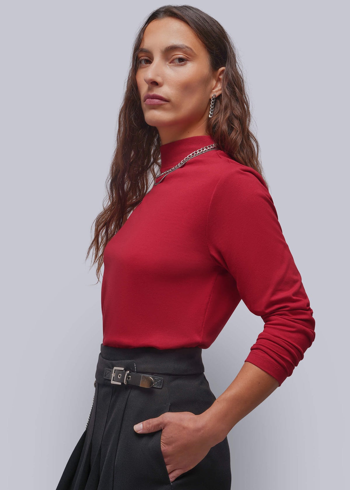 S=A woman modeling a fitted long-sleeve mock neck top in deep red, paired with a black skirt and a silver chain necklace, against a solid gray studio background.