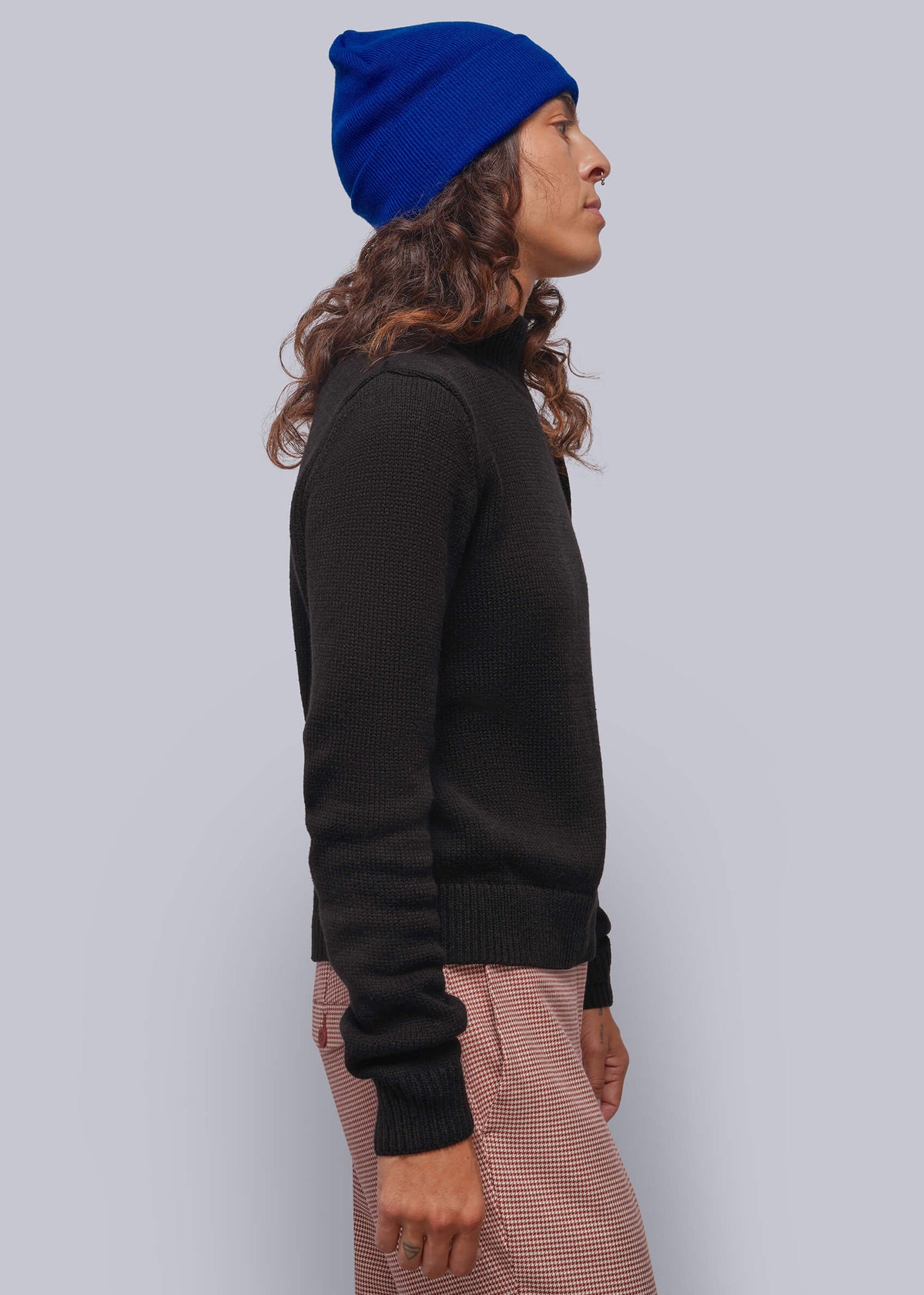 S=A side-profile view of a person wearing a bright royal blue knit beanie, a black long-sleeve knit sweater, and red-and-white houndstooth trousers, set against a solid light gray studio background.