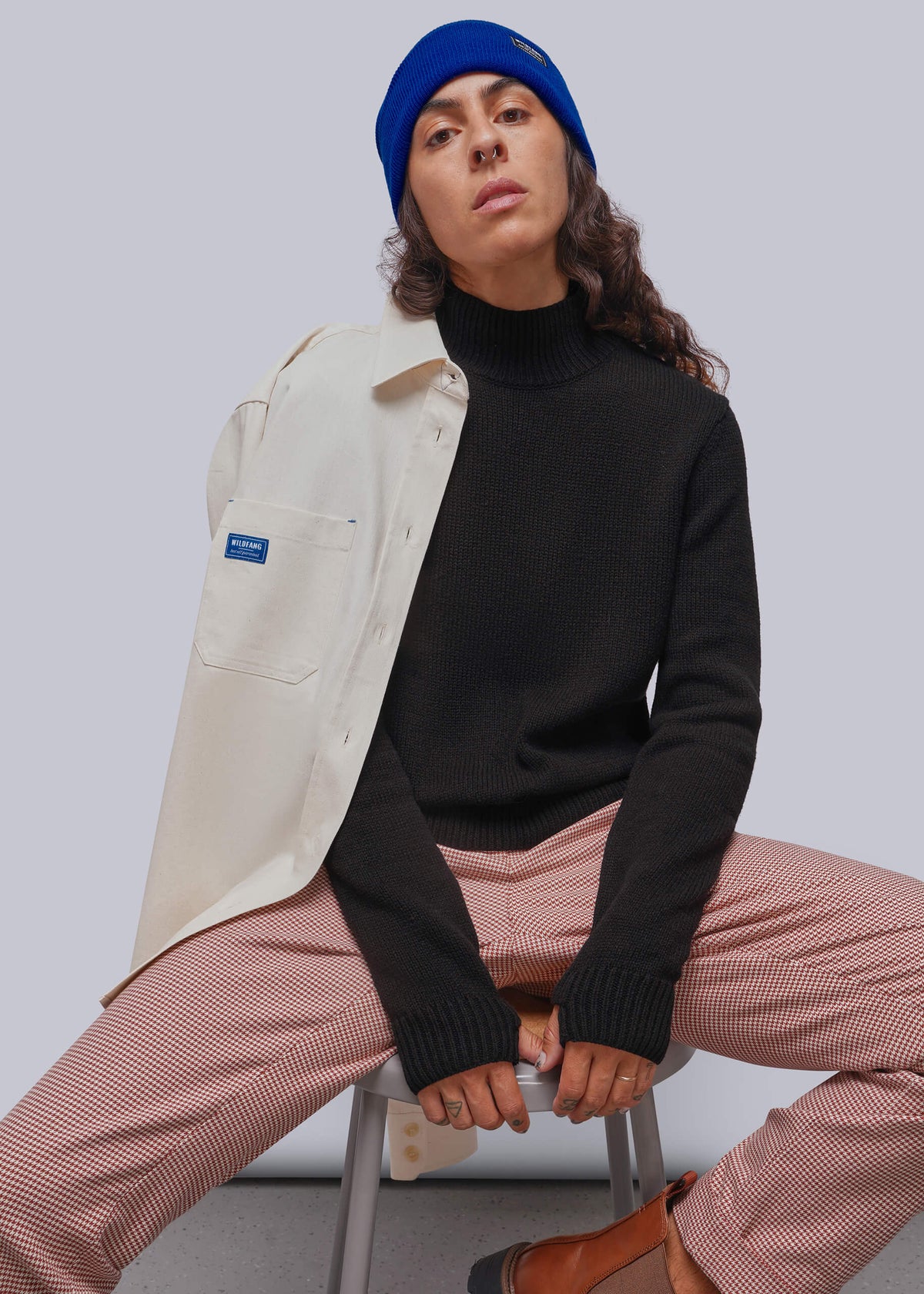 SA model wearing a black turtleneck sweater, red houndstooth trousers, a blue beanie, and a cream-colored button up draped off their shoulder, seated on a stool against a light gray background.