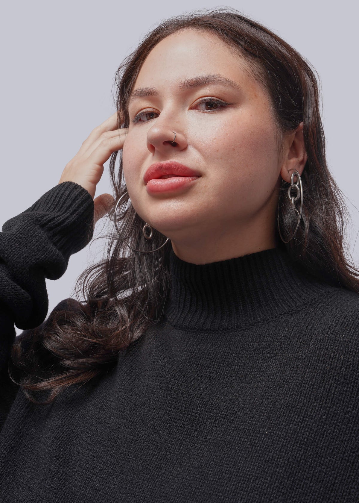 1X=A close-up portrait of a woman wearing a black ribbed turtleneck sweater and silver hoop earrings against a light gray studio background.