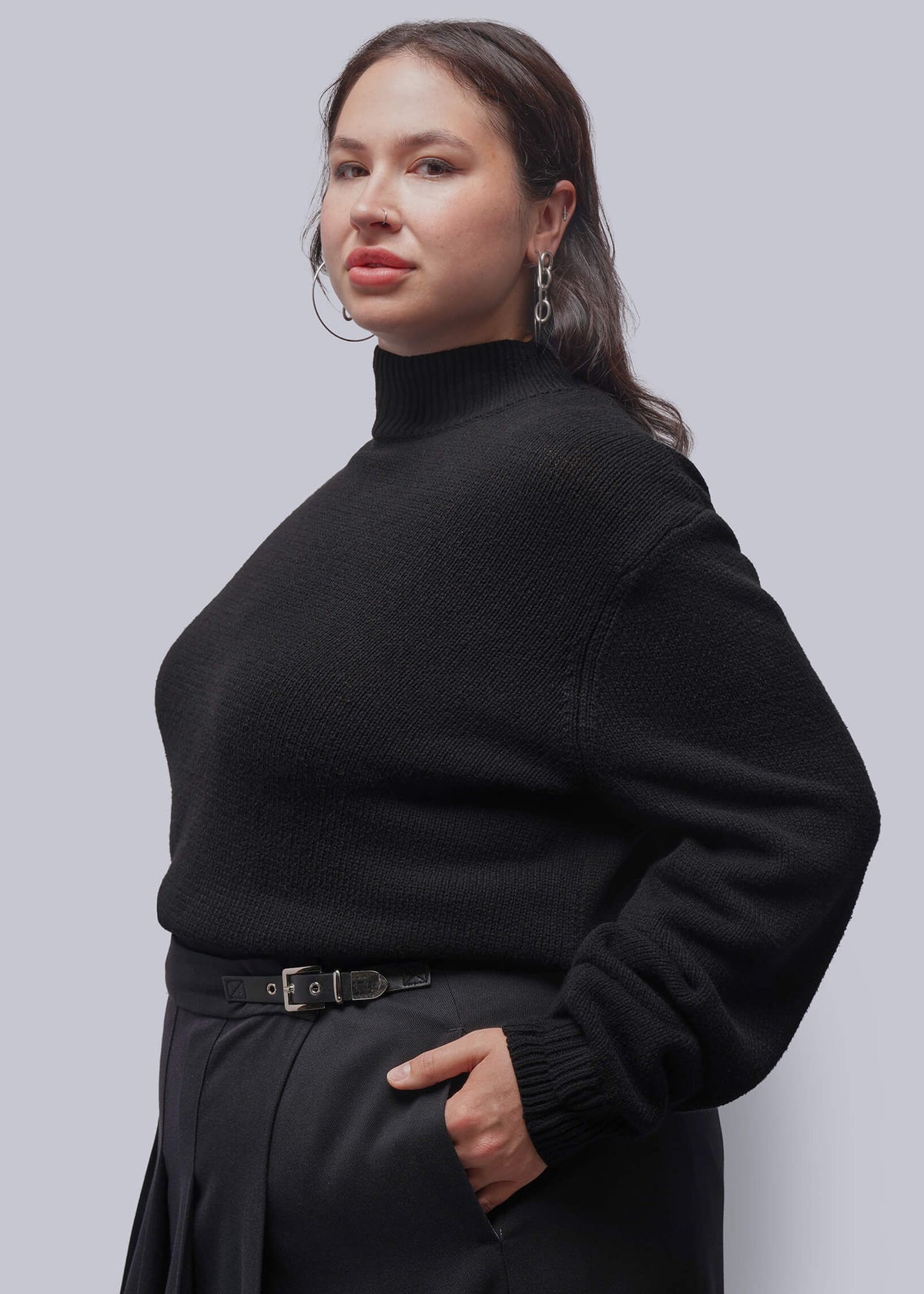 1X=A woman wearing a black ribbed turtleneck sweater and black skirt, posing against a solid gray studio background.