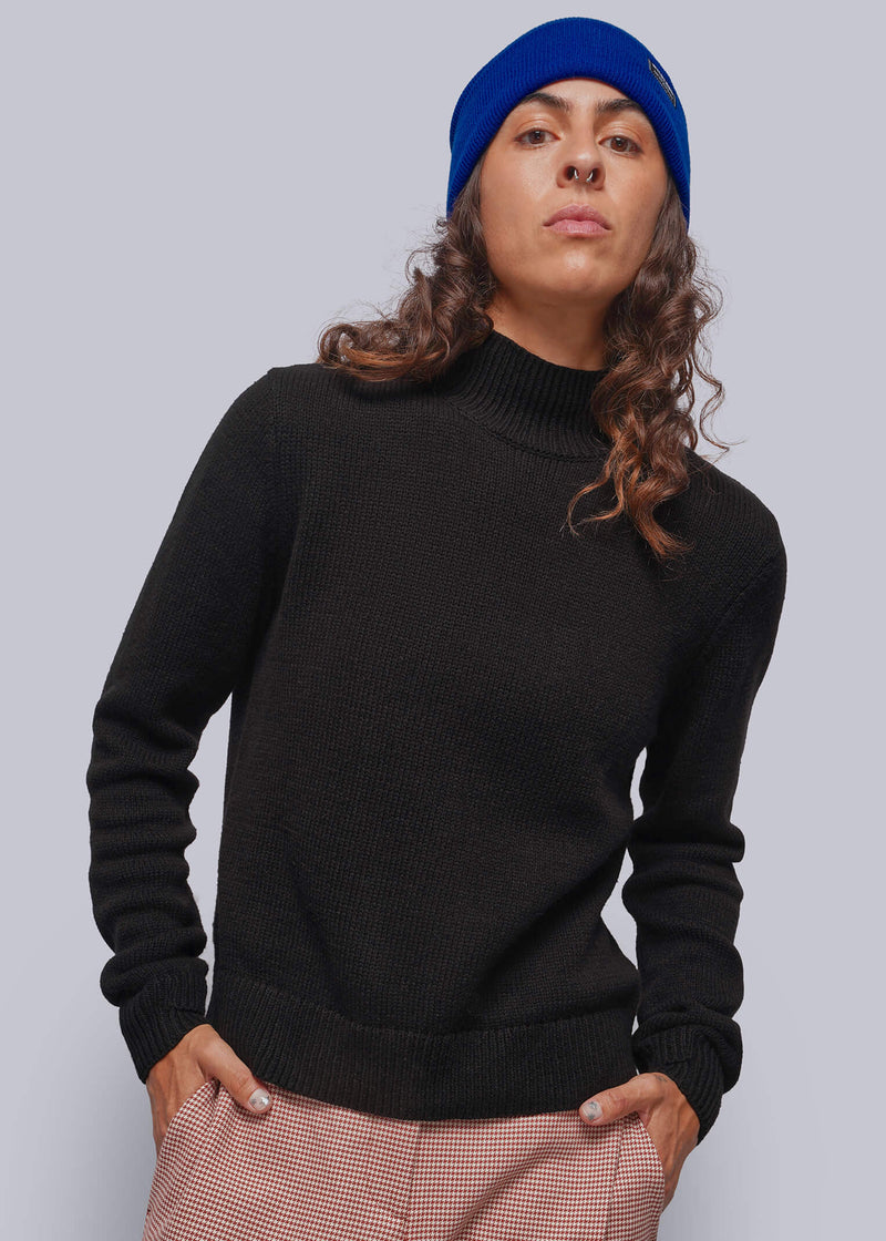S=A person wearing a black mock neck knit sweater, red patterned houndstooth trousers, and a bright blue beanie, standing against a solid gray studio background.