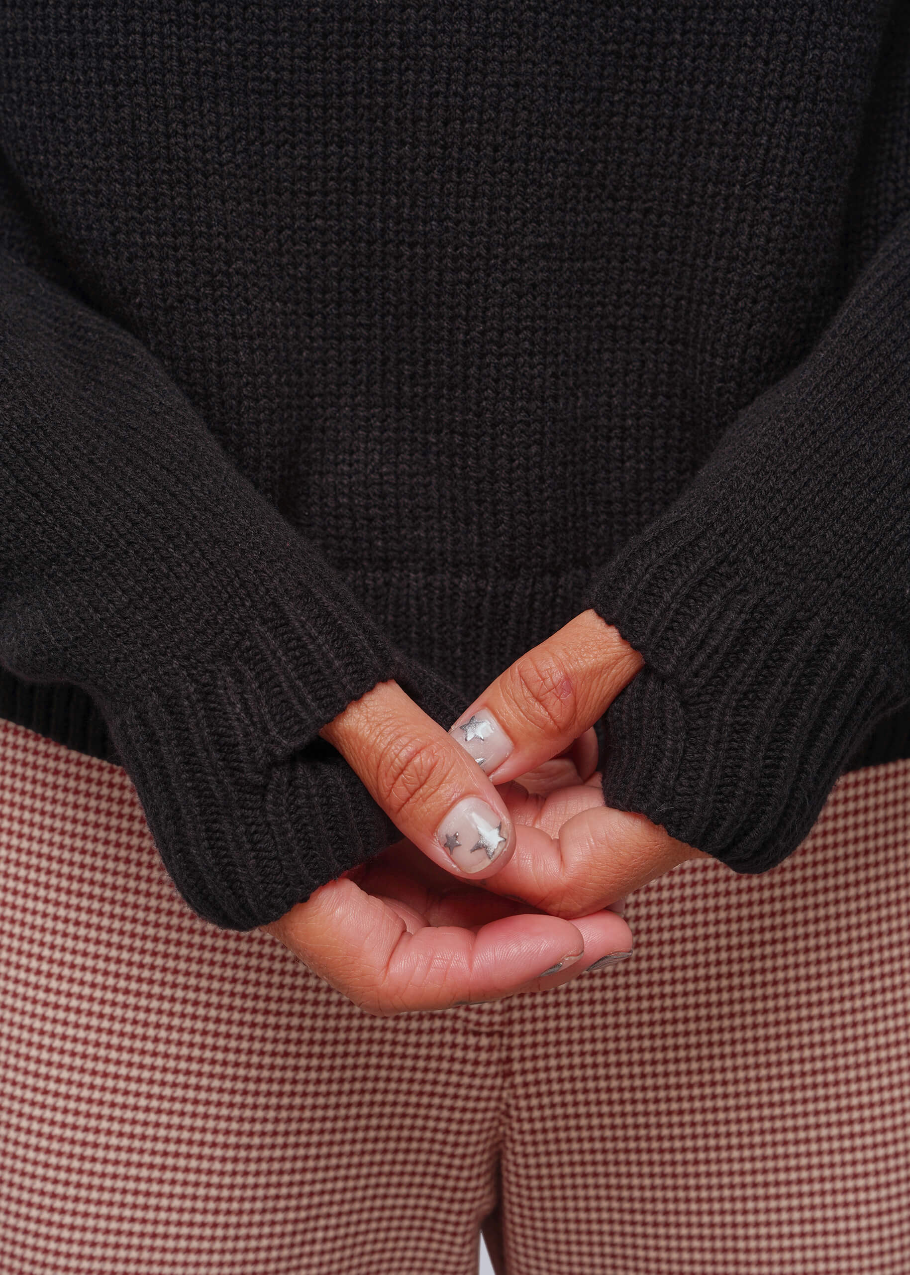 Close-up of a person wearing a black ribbed knit sweater with thumbholes and houndstooth trousers, with hands clasped in front.