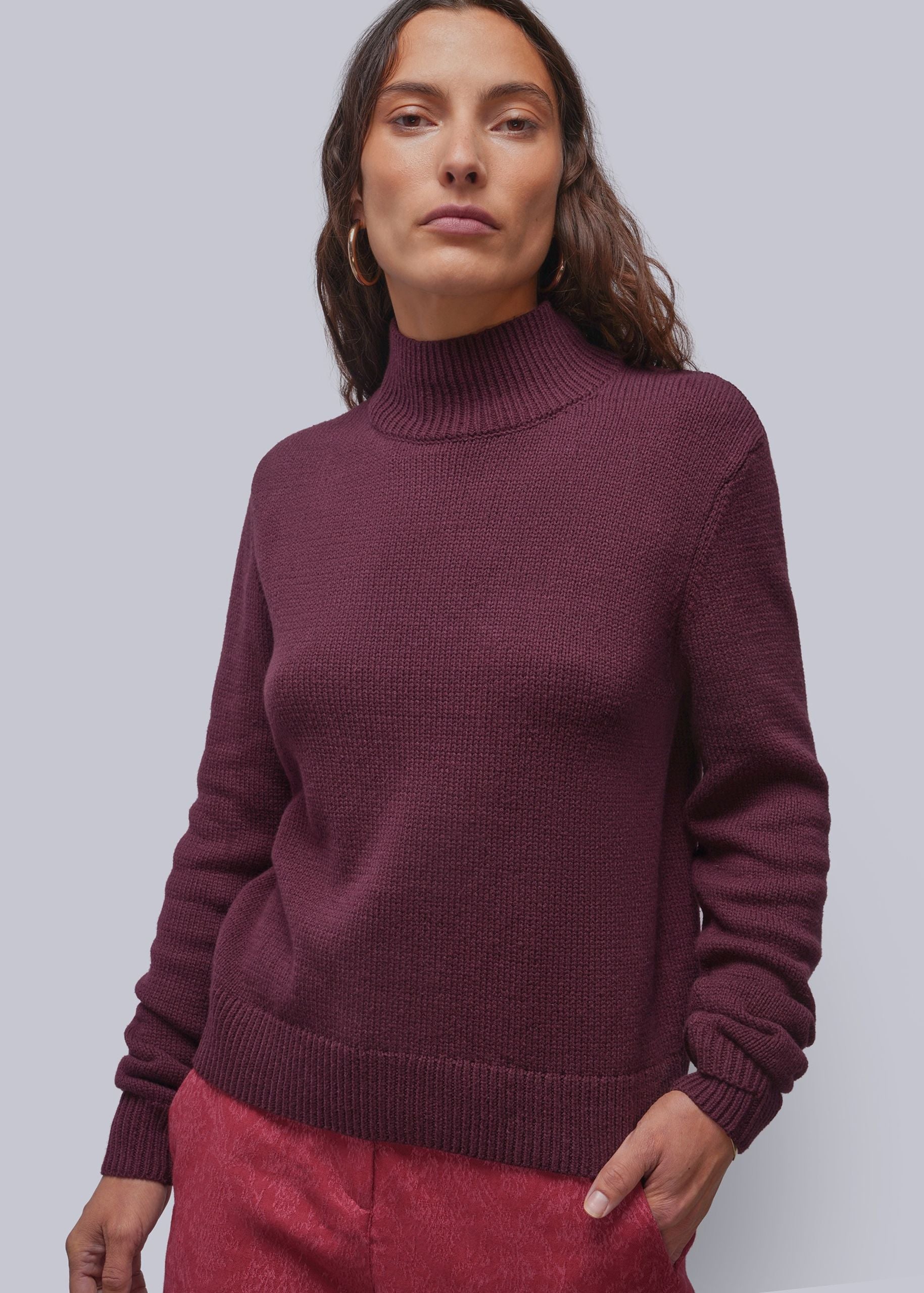 A woman wearing a ribbed turtleneck sweater in a deep burgundy color, paired with textured pink pants, against a solid light gray background.