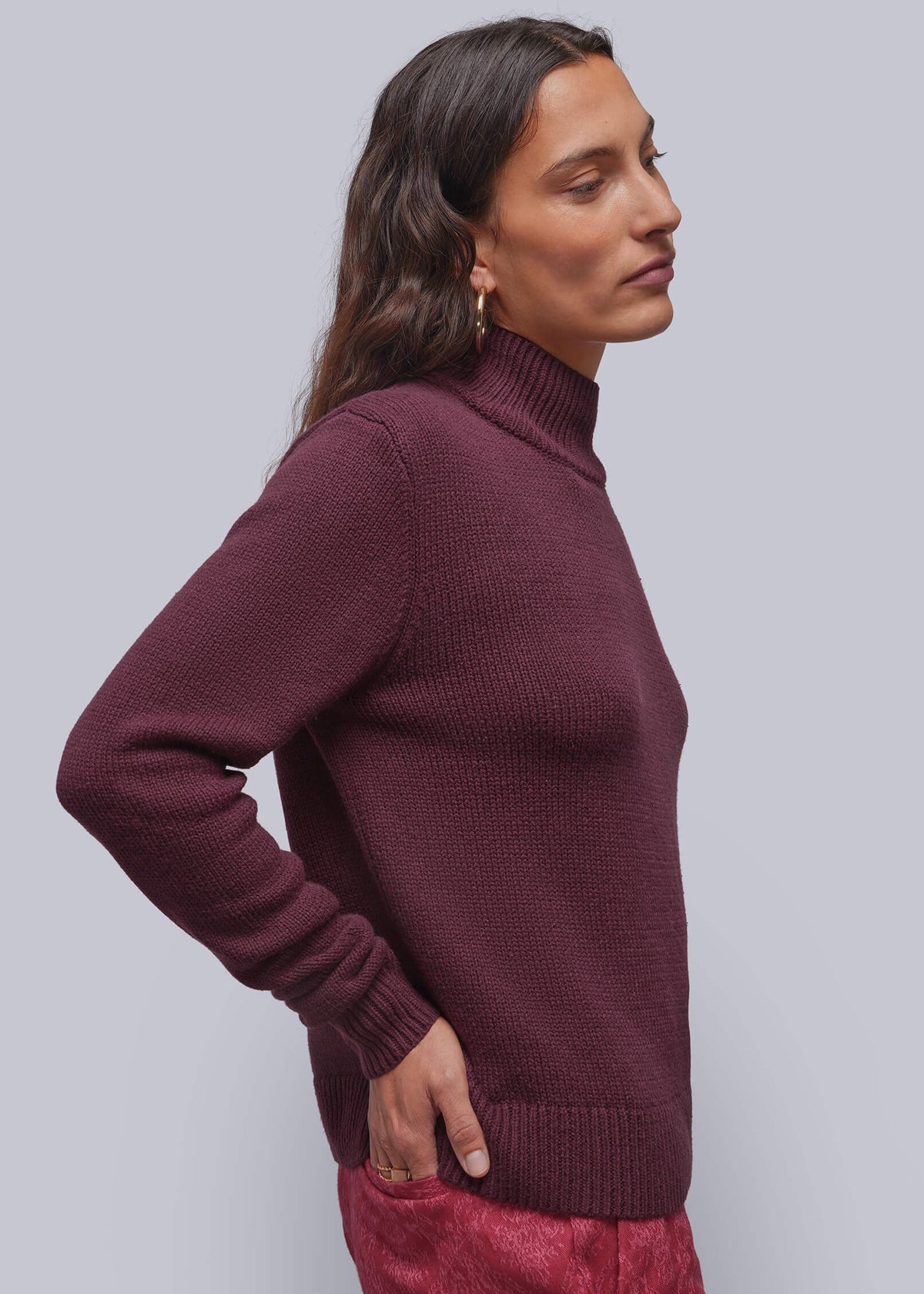 S=A woman in profile wearing a ribbed turtleneck sweater in deep burgundy and matching textured trousers, set against a solid light gray studio background.