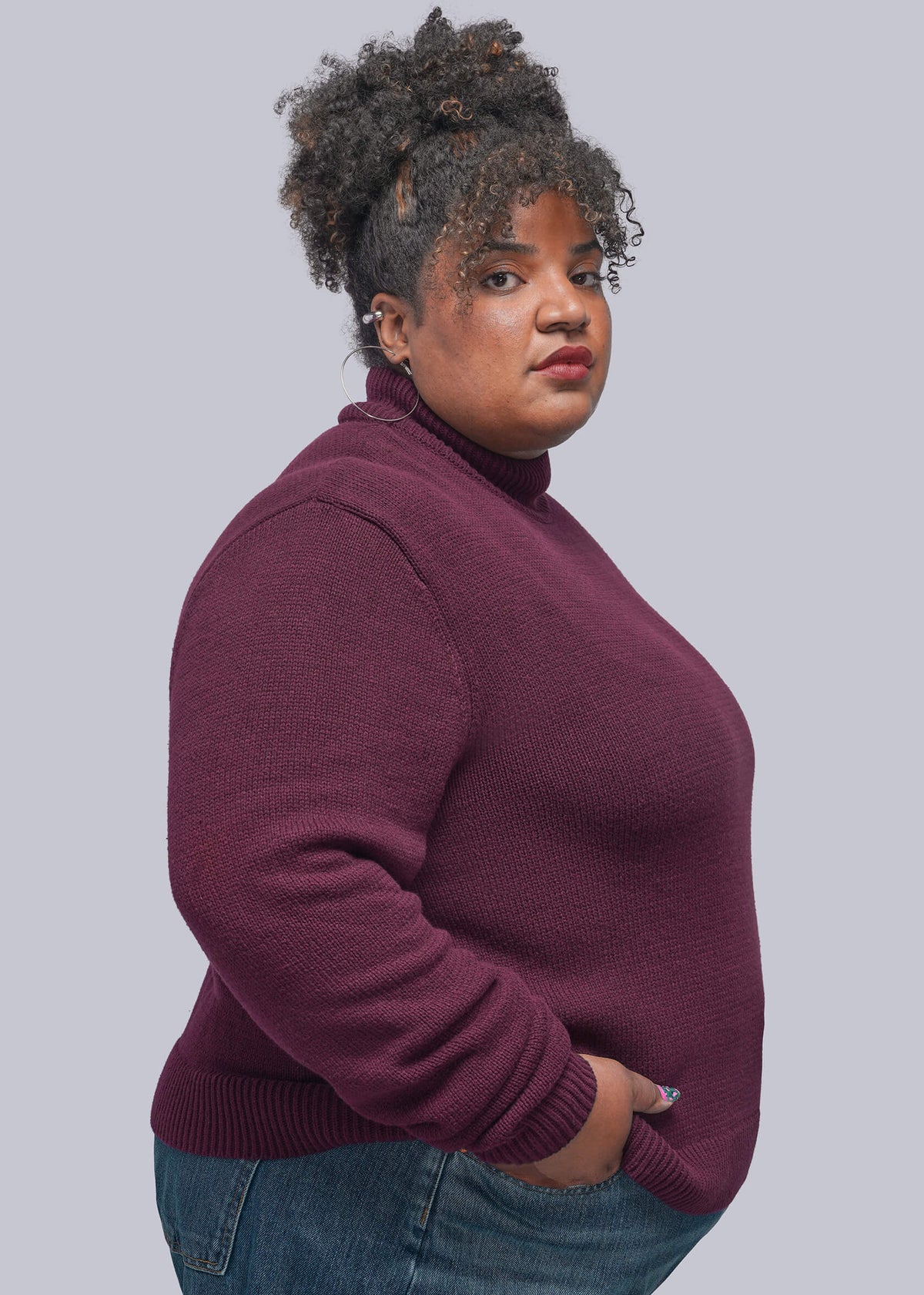 1X=A woman with curly hair wearing a ribbed turtleneck sweater in deep burgundy and blue jeans, standing against a solid gray background.