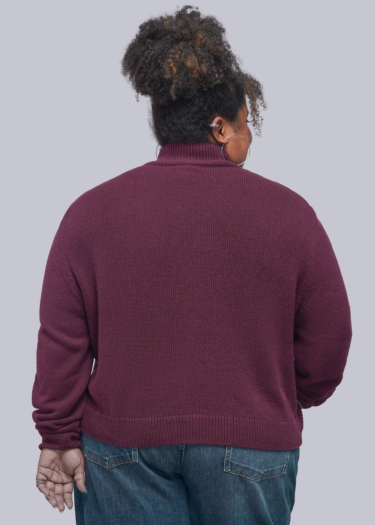 1X=A person shown from the back wearing a ribbed turtleneck sweater in a deep burgundy or plum shade, paired with blue jeans, against a solid light gray studio background.