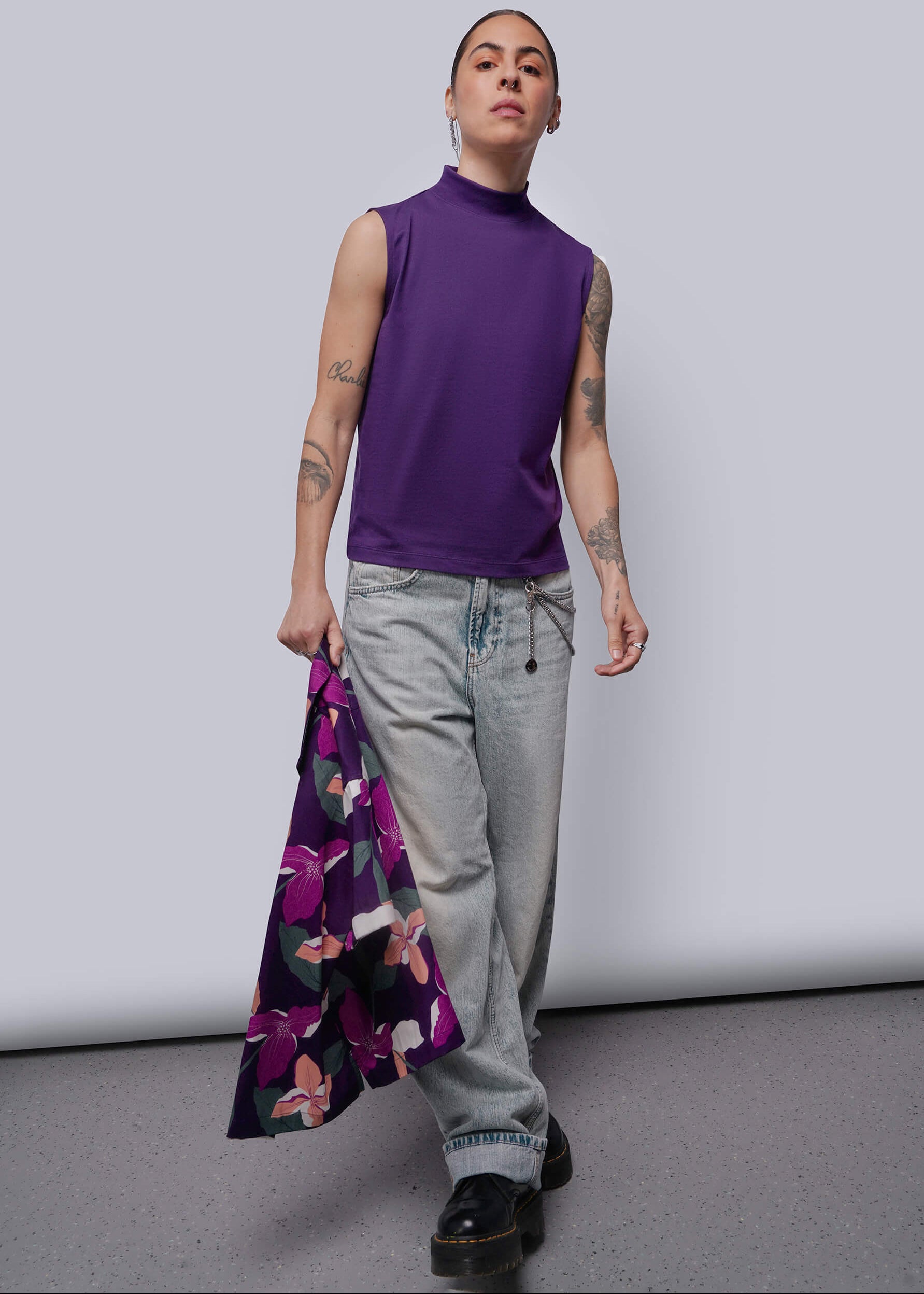 S=Person wearing a purple sleeveless top and gray pants, holding a floral shirt against a plain background.