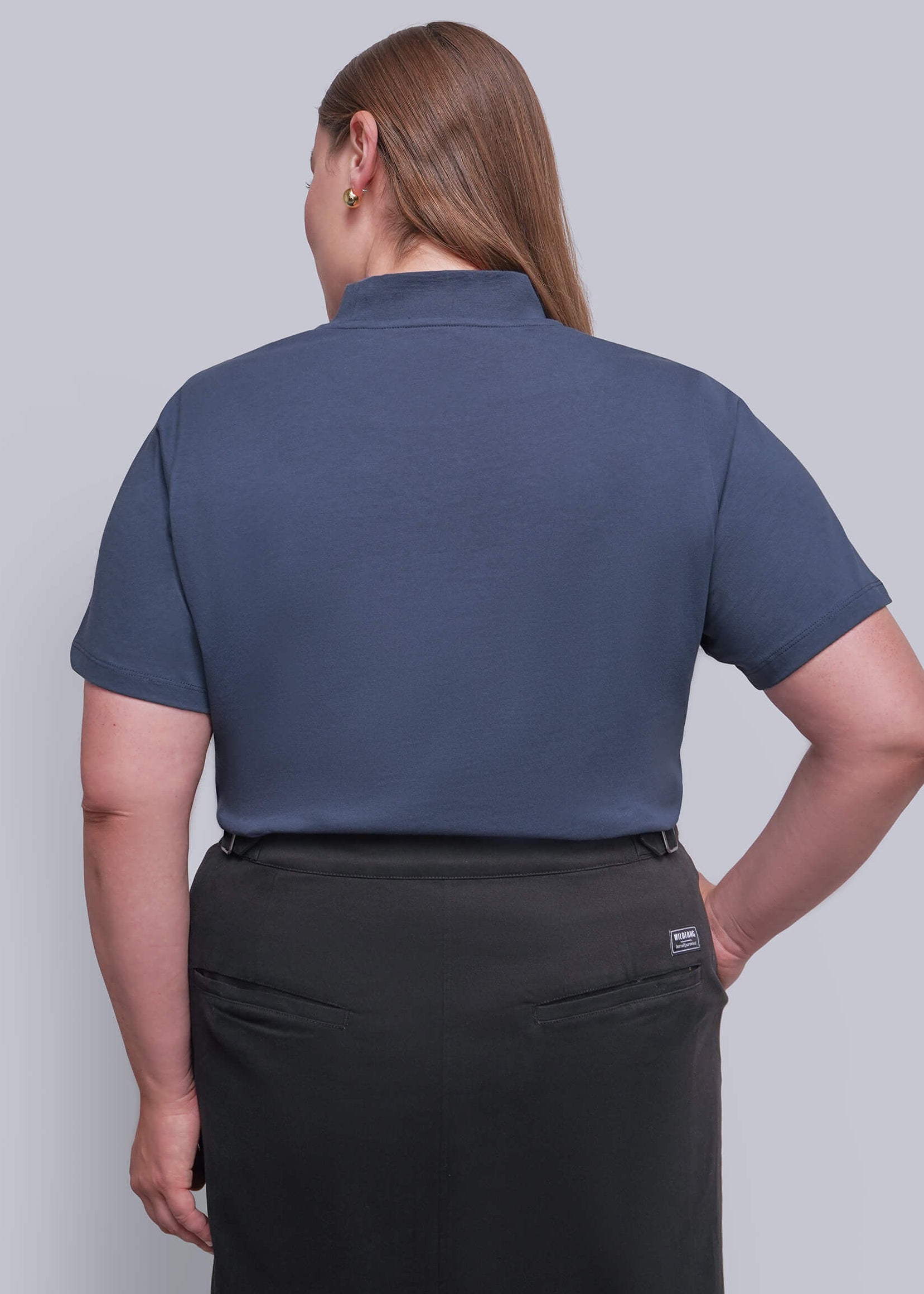 1X=A woman shown from the back wearing a slate blue short-sleeve polo shirt and a black skirt, photographed against a solid light gray studio background.