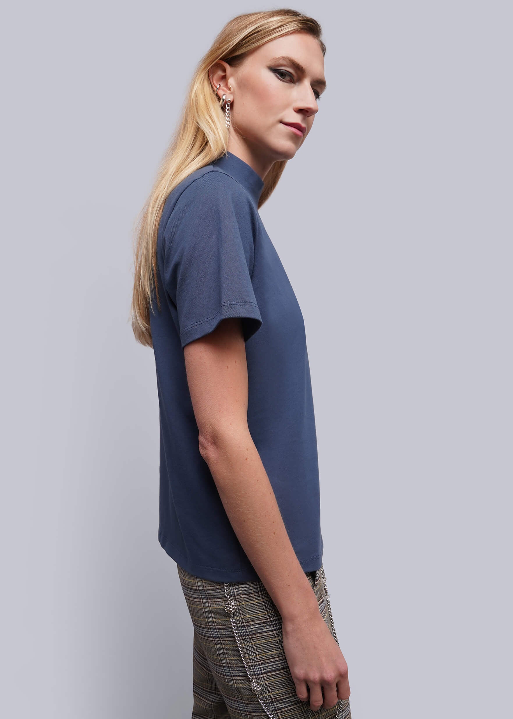 S=A woman in side profile wearing a short-sleeve blue mock-neck top and plaid trousers, photographed against a solid light gray studio background.