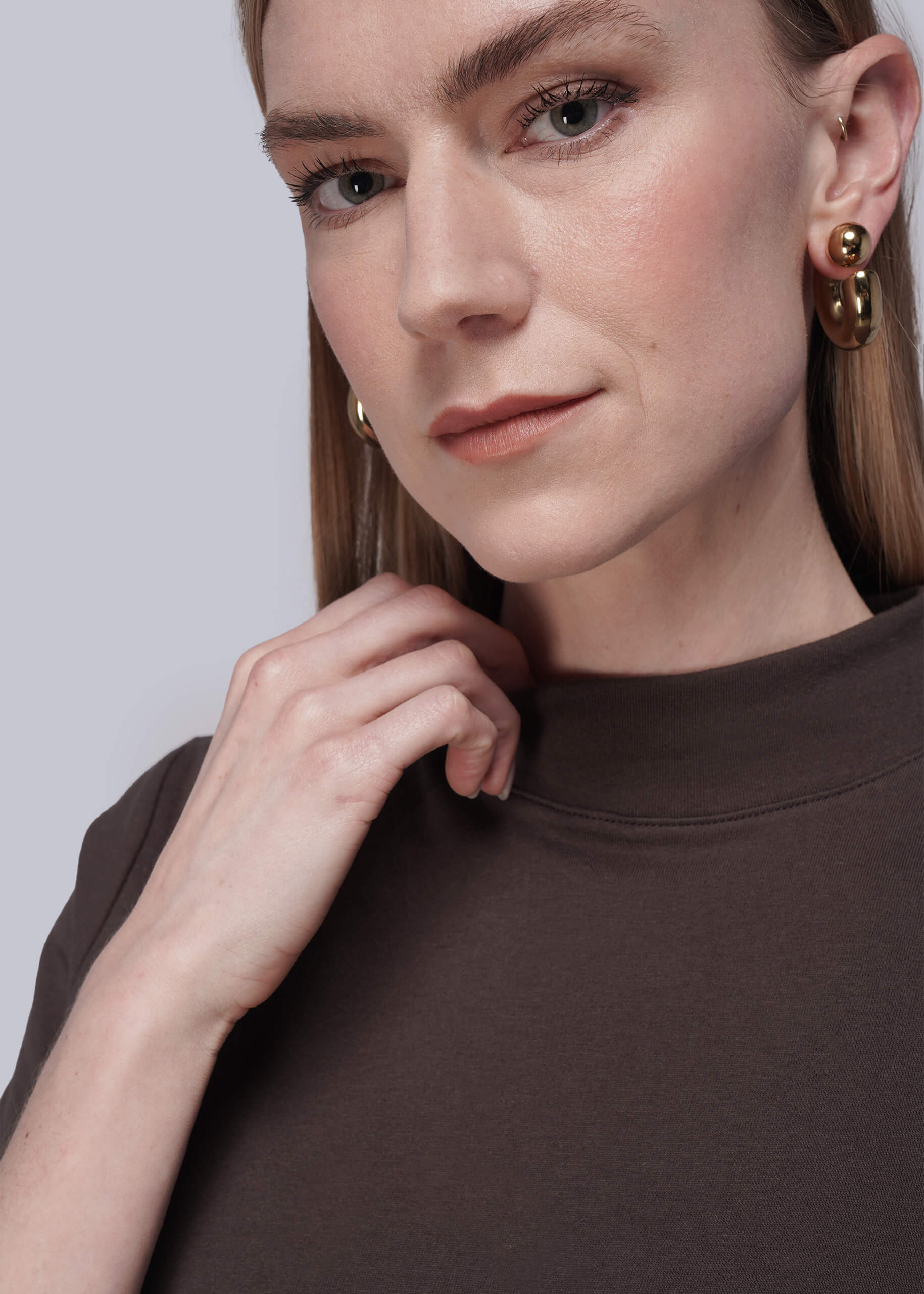 Close-up of a person wearing brown shirt and gold earrings against a light gray background