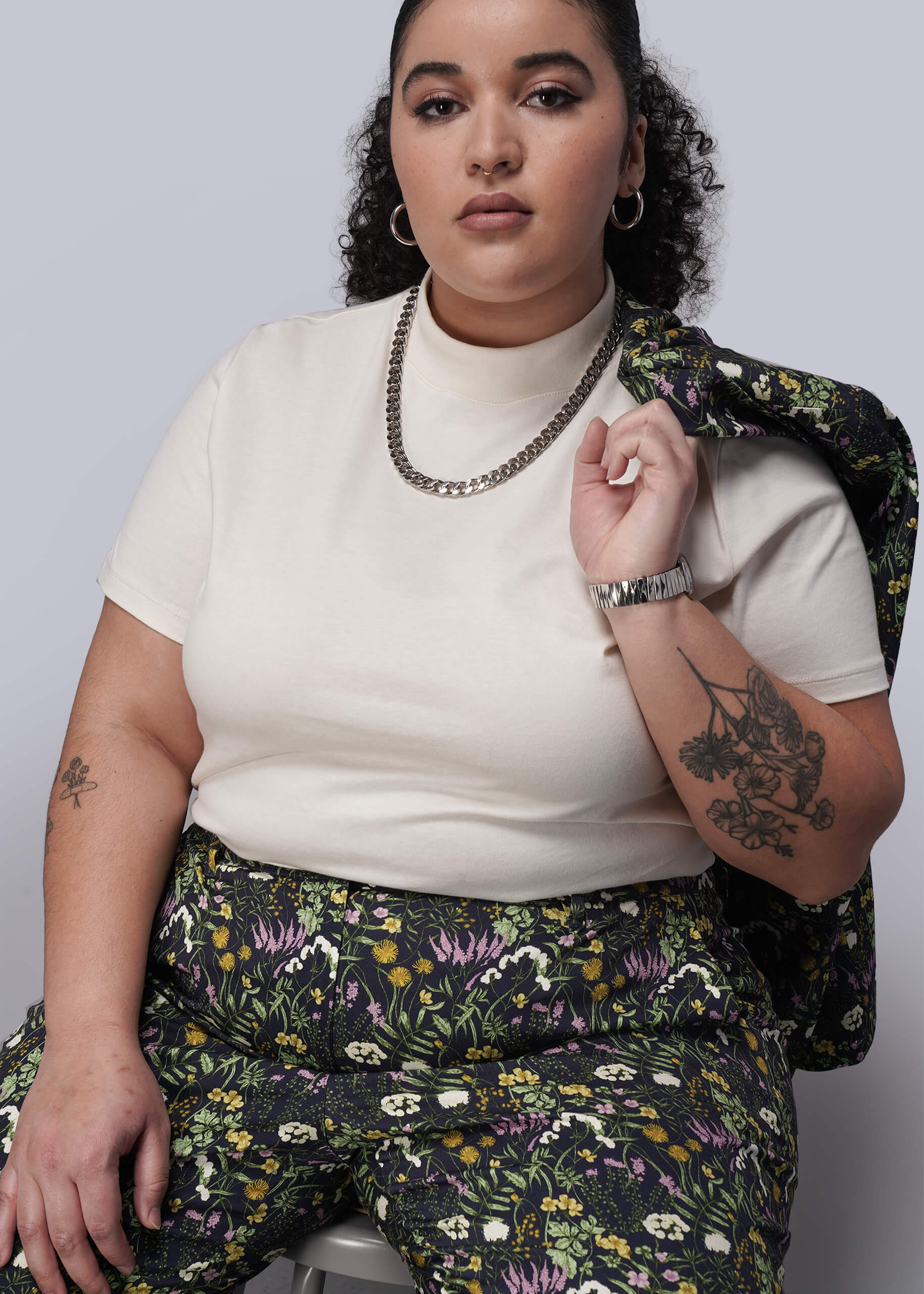 1X=Woman wearing a white top and dark floral pants against a plain background