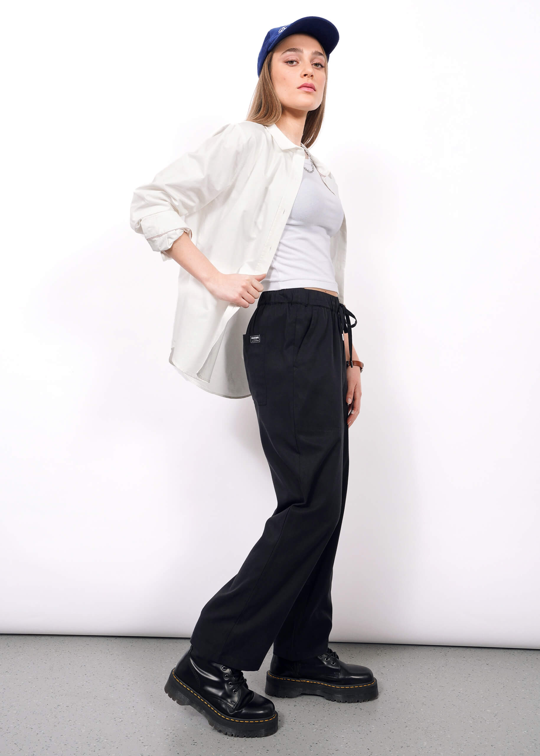 Wearing The Everyday Elastic Waist Pant in black, paired with a blue cap, white shirt over a white tank top, and black platform shoes, a person stands against a plain white background and looks confidently at the camera.