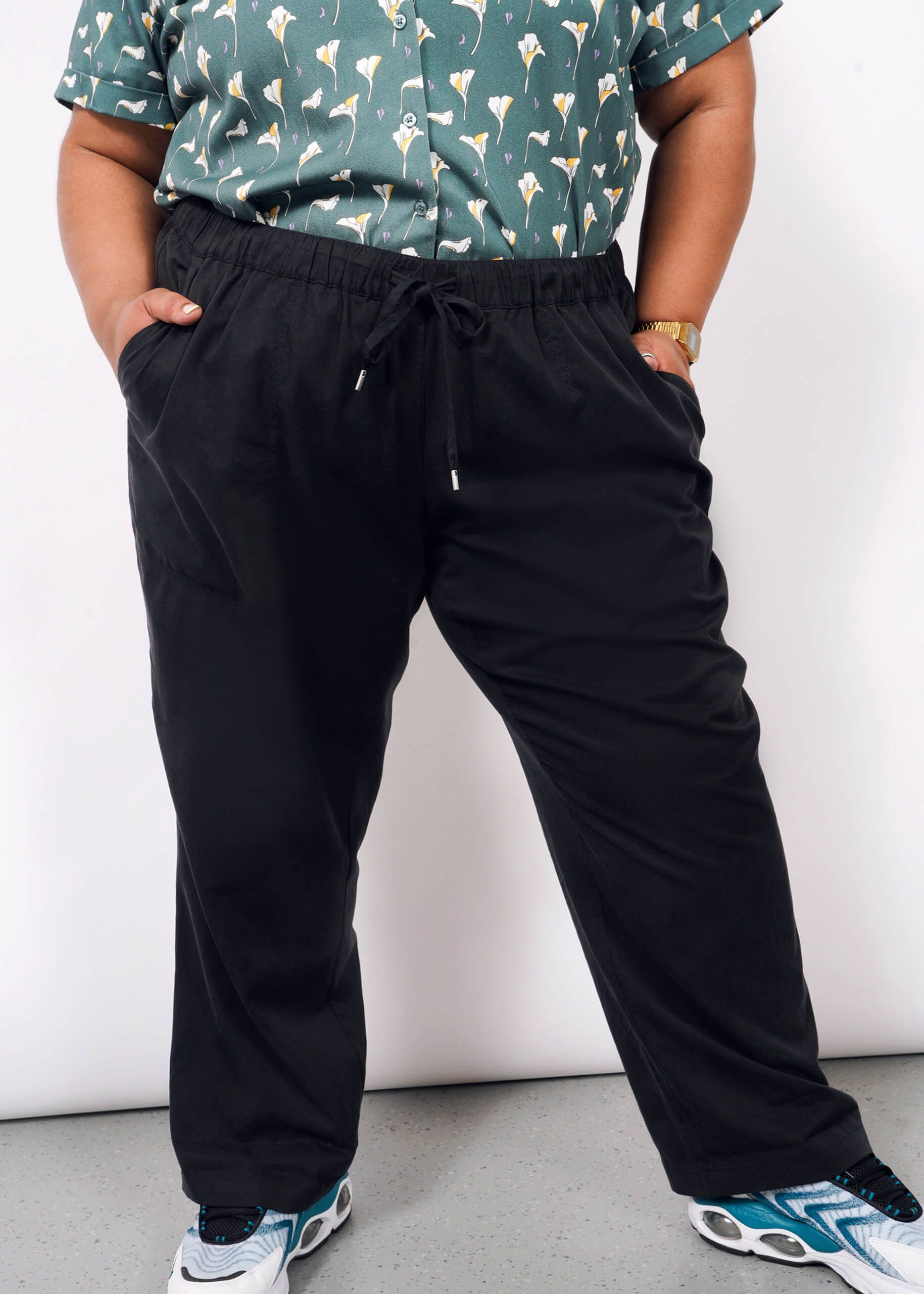 A person stands with hands in their pockets against a plain white background, wearing a green patterned shirt, The Everyday Elastic Waist Pant in black, white and blue sneakers, and a gold watch.