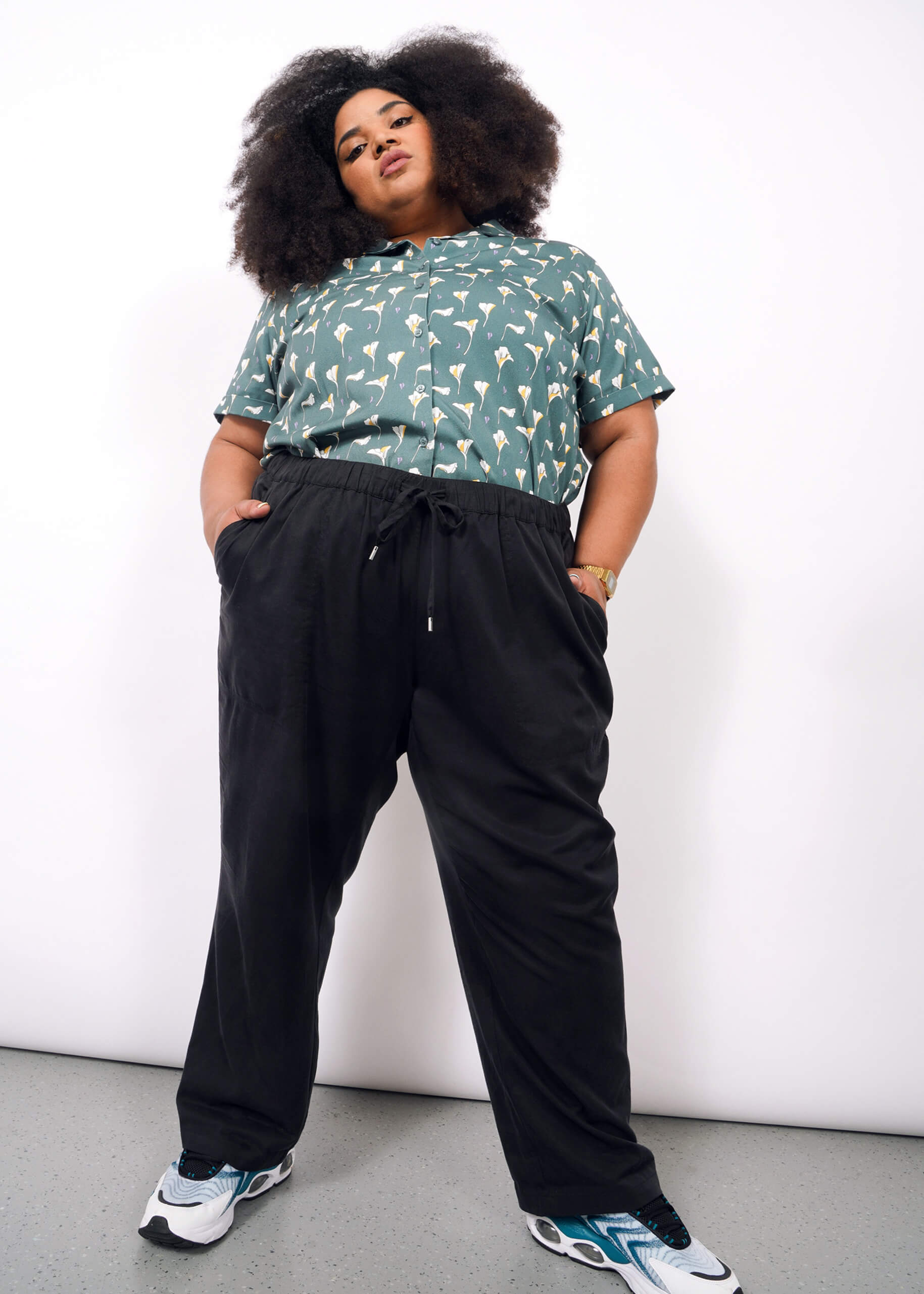 A person with voluminous curly hair stands confidently against a white background, wearing a green patterned short-sleeve shirt, The Everyday Elastic Waist Pant in black, a watch, and white, black, and teal sneakers.