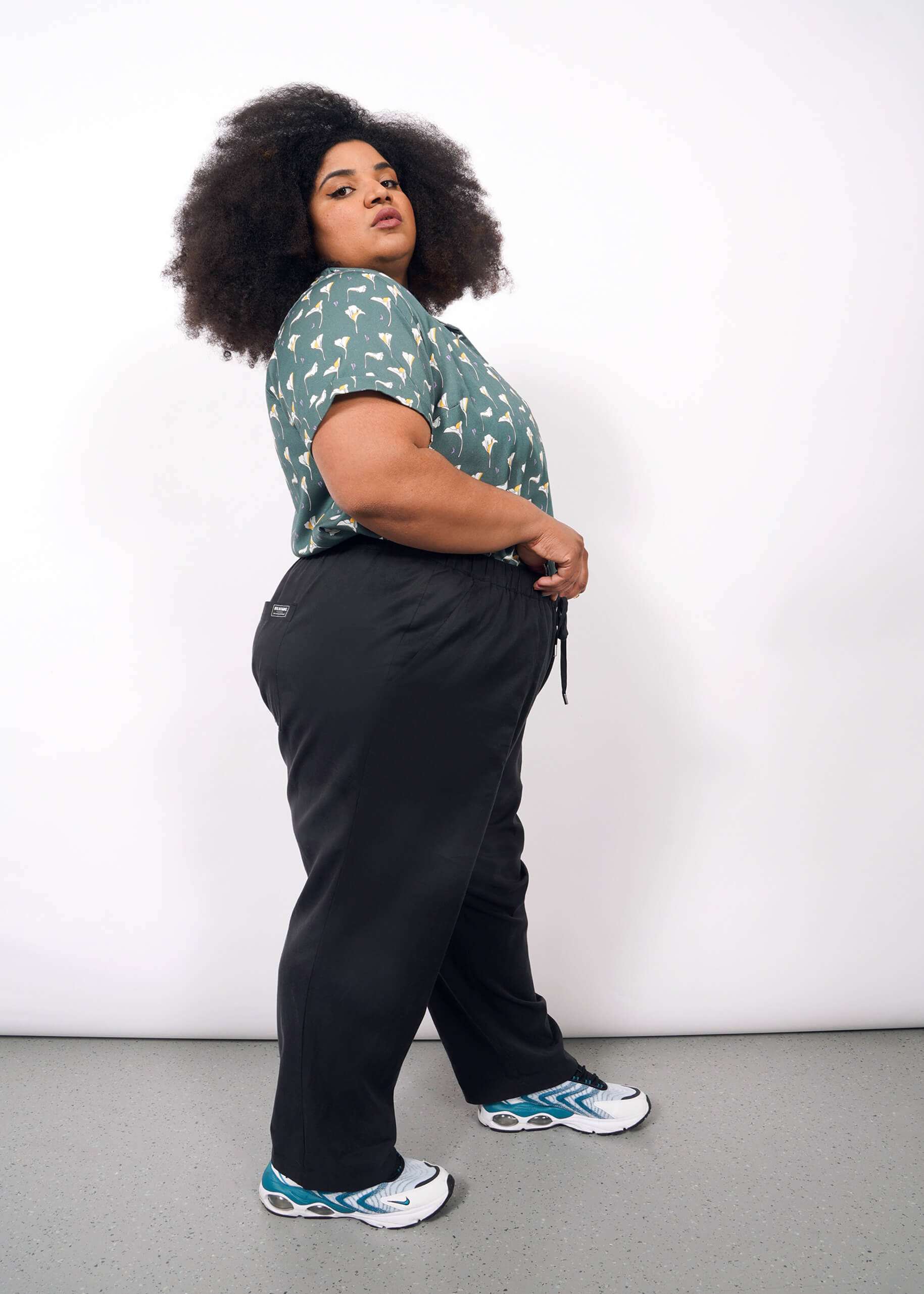 A person with curly hair stands sideways against a white background, wearing a green patterned shirt, The Everyday Elastic Waist Pant in black, and white sneakers with blue accents.
