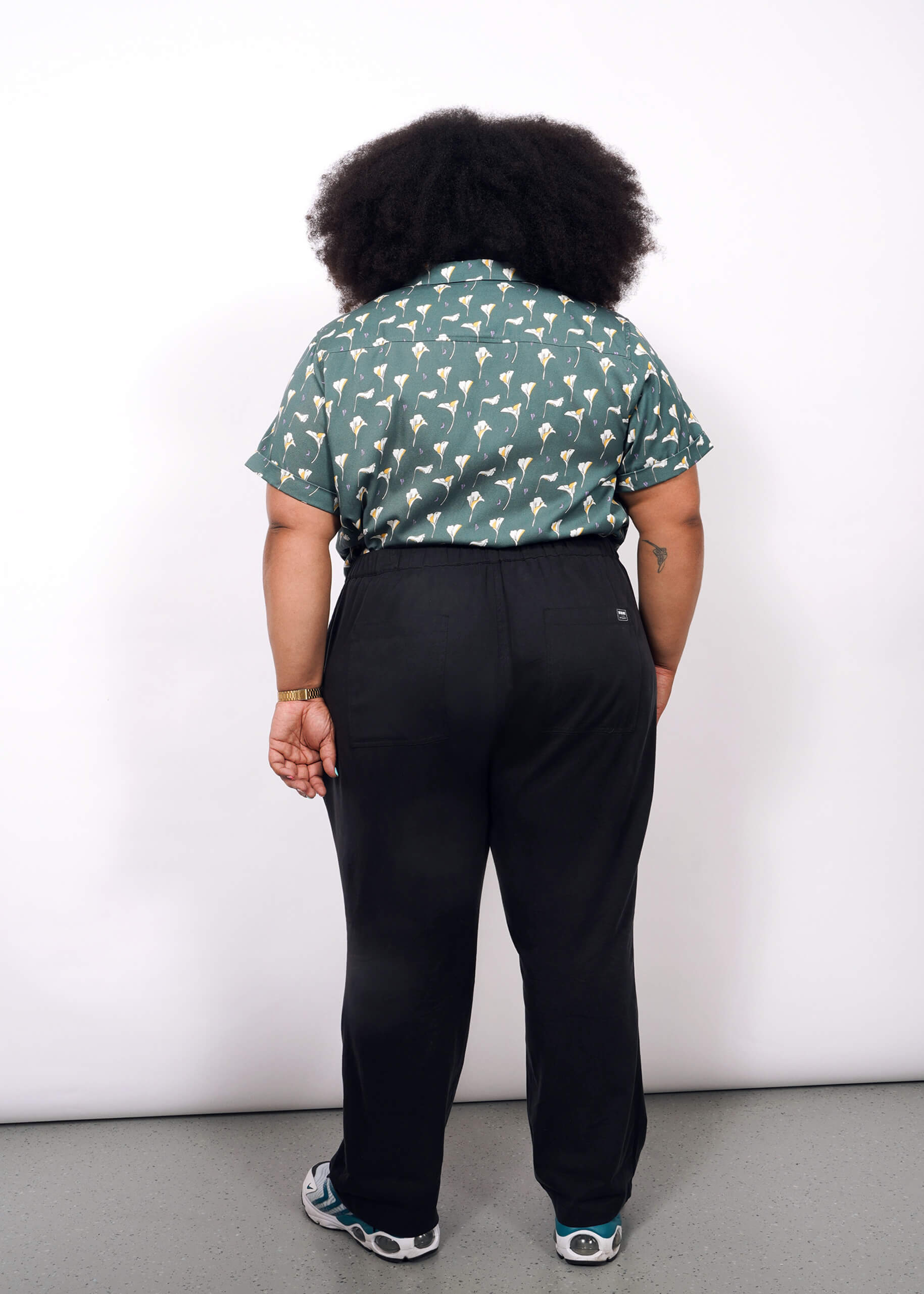 A person with curly hair faces away from the camera, wearing a green patterned short-sleeve shirt, The Everyday Elastic Waist Pant in black, and sneakers, set against a plain white background.