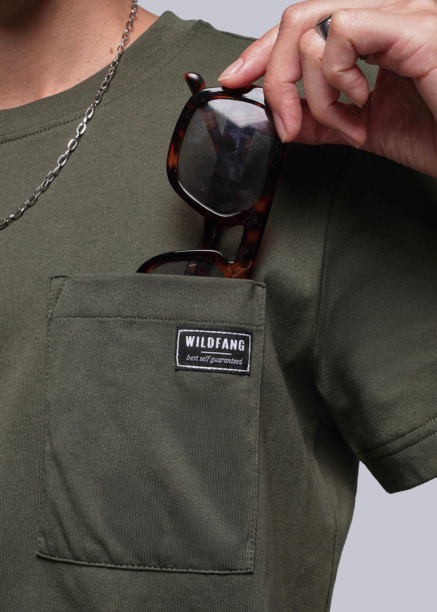 Person wearing a green t-shirt with a Wildfang logo on pocket, holding sunglasses.