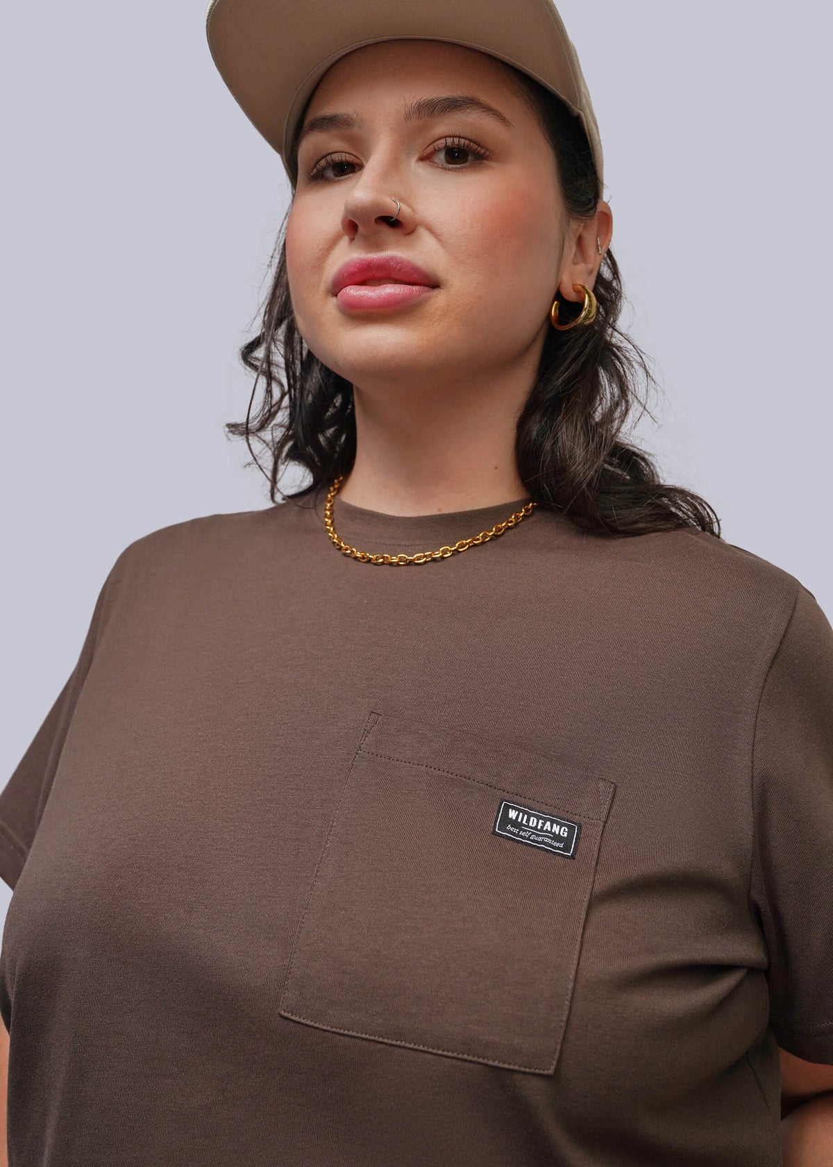 1X=A woman wearing a brown short-sleeve crewneck t-shirt with a chest pocket and a small black 'WILDFANG' brand label, paired with a cap, against a solid gray background.