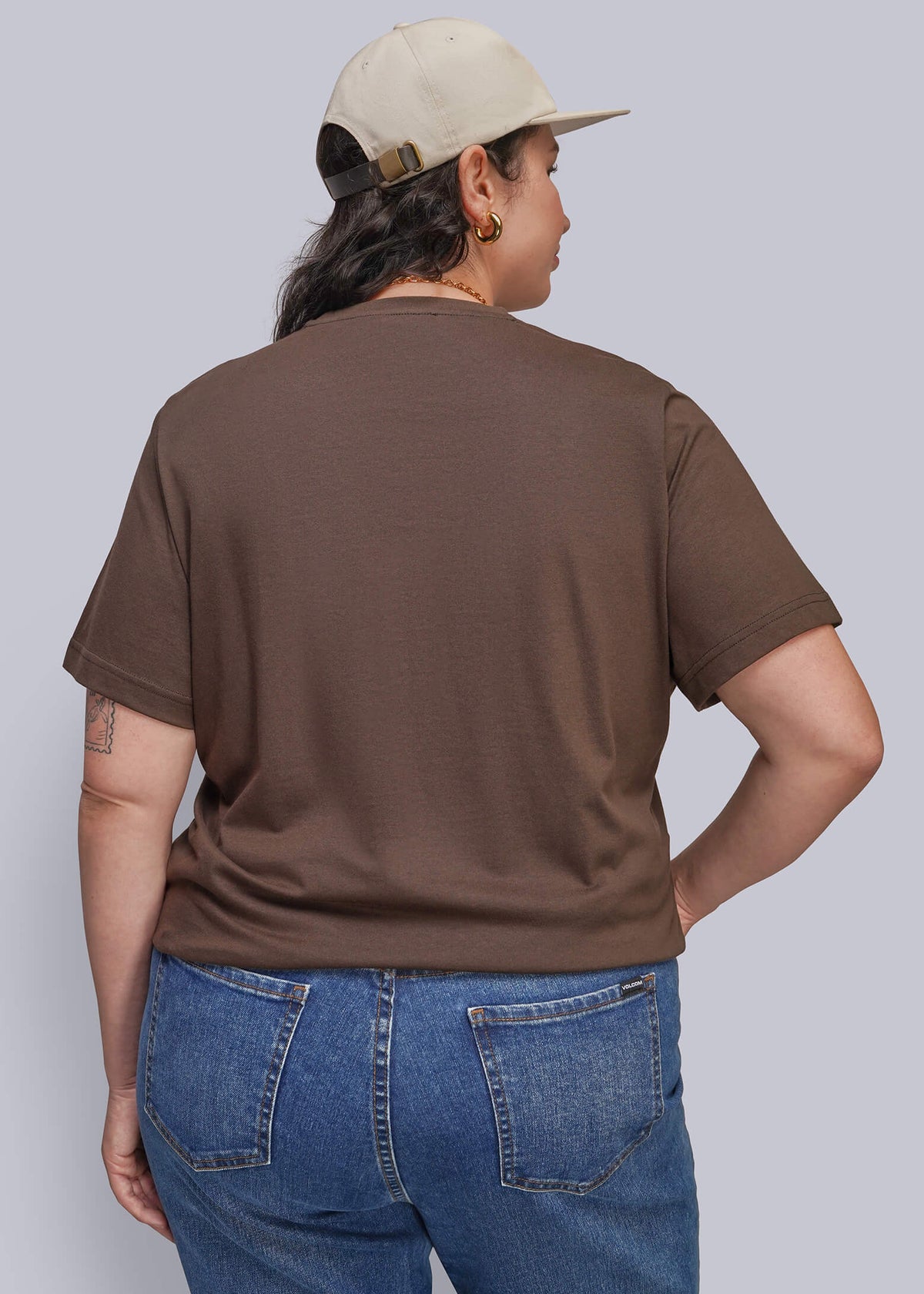 1X=A person shown from the back wearing a brown short-sleeve crewneck t-shirt, blue jeans, and a beige baseball cap, against a solid light gray background.