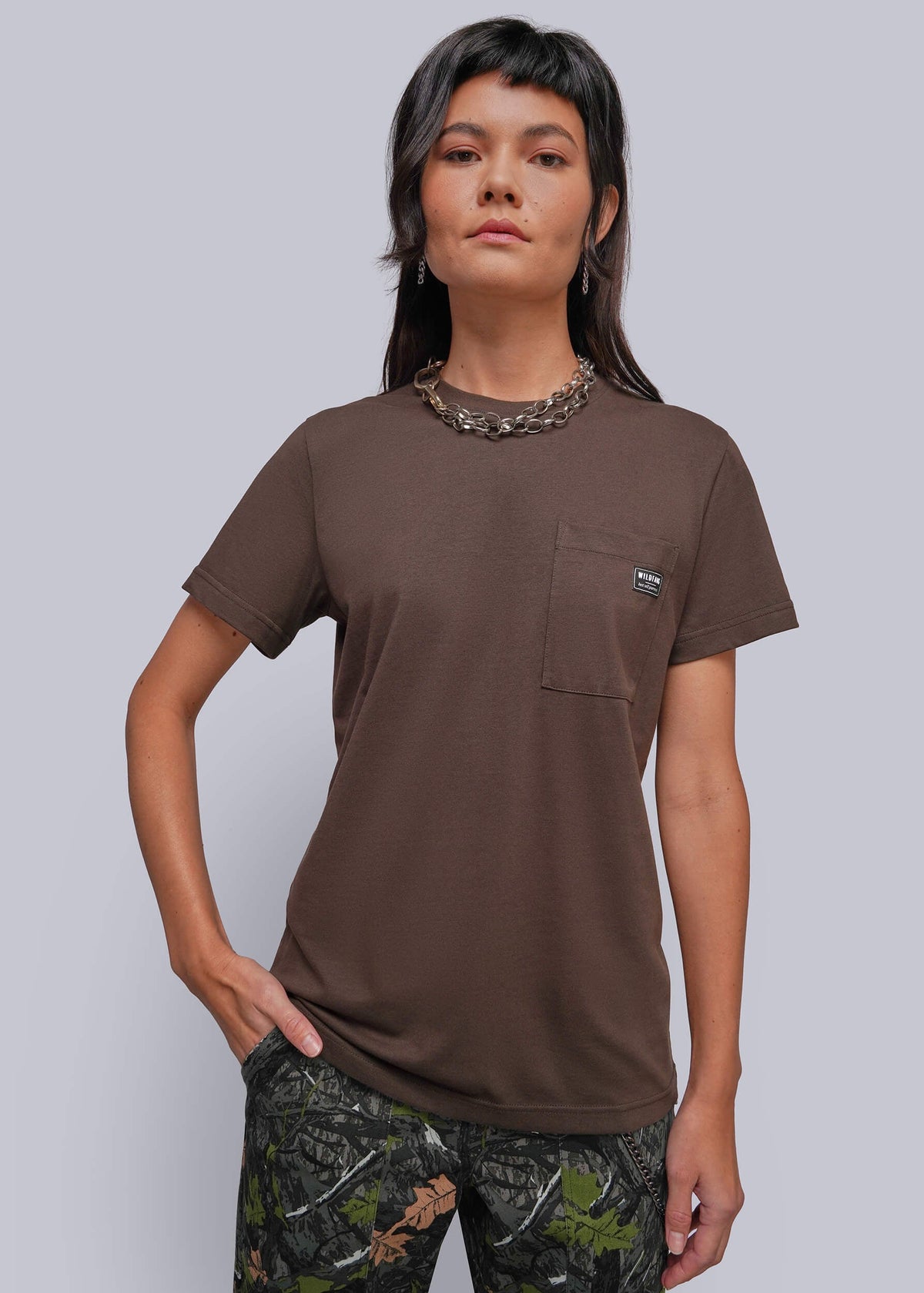 S=A woman modeling a brown short-sleeve pocket t-shirt with a small embroidered logo on the chest pocket, paired with camouflage pants, against a solid gray studio background.