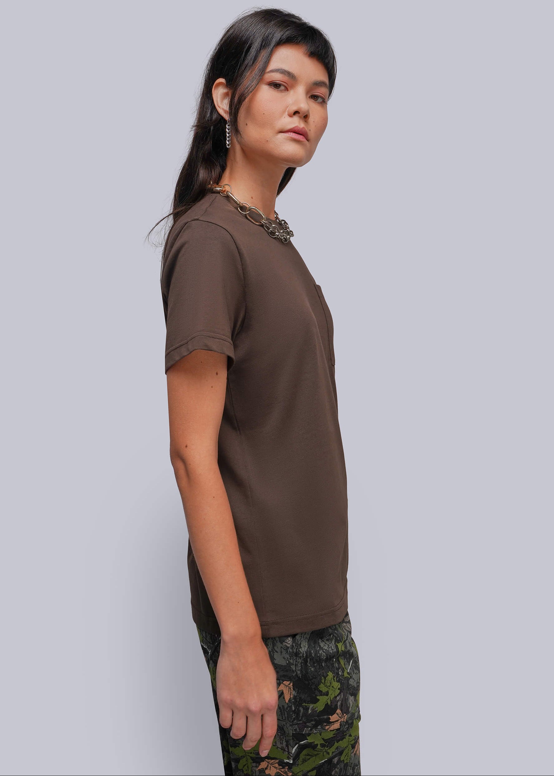 A woman wearing a brown short-sleeve crewneck t-shirt and a dark green floral print skirt, standing against a solid light gray background.