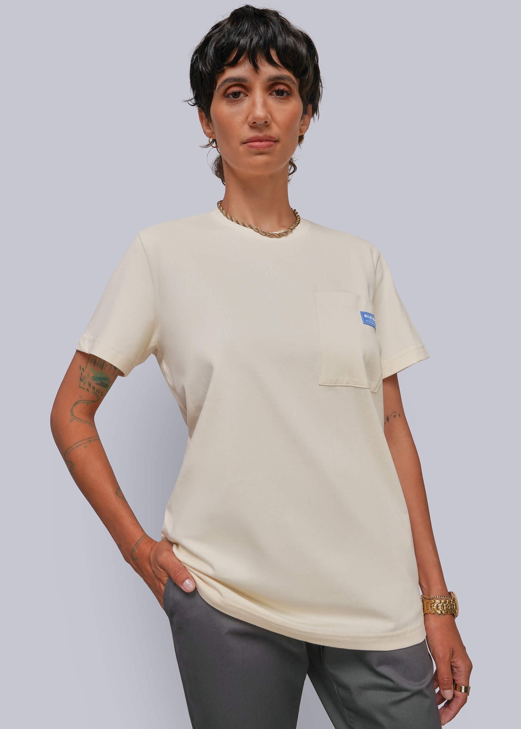 A model wearing a cream-colored short-sleeve pocket t-shirt with a small blue logo on the chest pocket, paired with gray pants, against a solid light gray background.