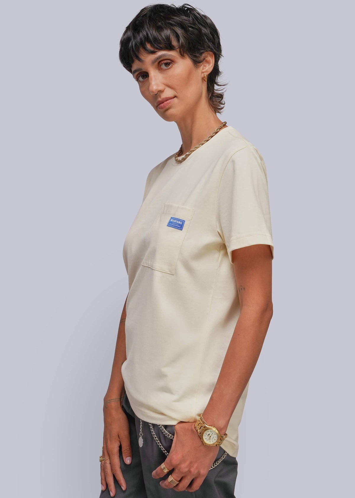 S=A person modeling a cream-colored short-sleeve pocket t-shirt with a small blue rectangular patch on the chest pocket, paired with dark gray pants, against a solid light gray background.