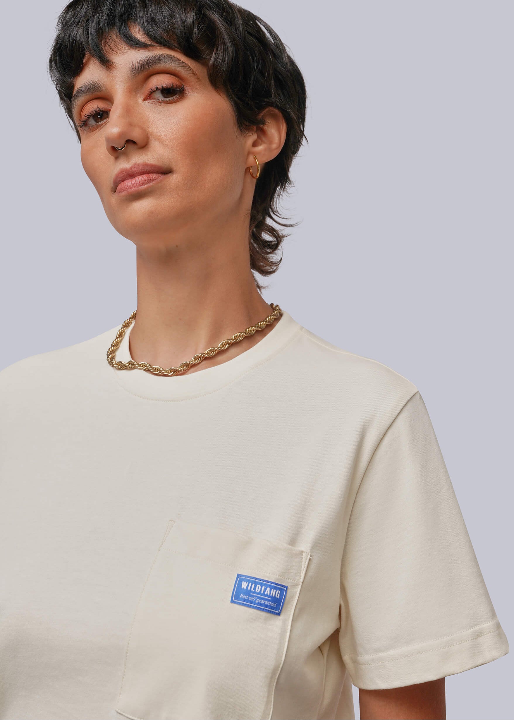 A close-up of a model wearing a cream-colored short-sleeve pocket t-shirt with a blue 'MIRIAM' brand label on the chest pocket, set against a solid light gray background.