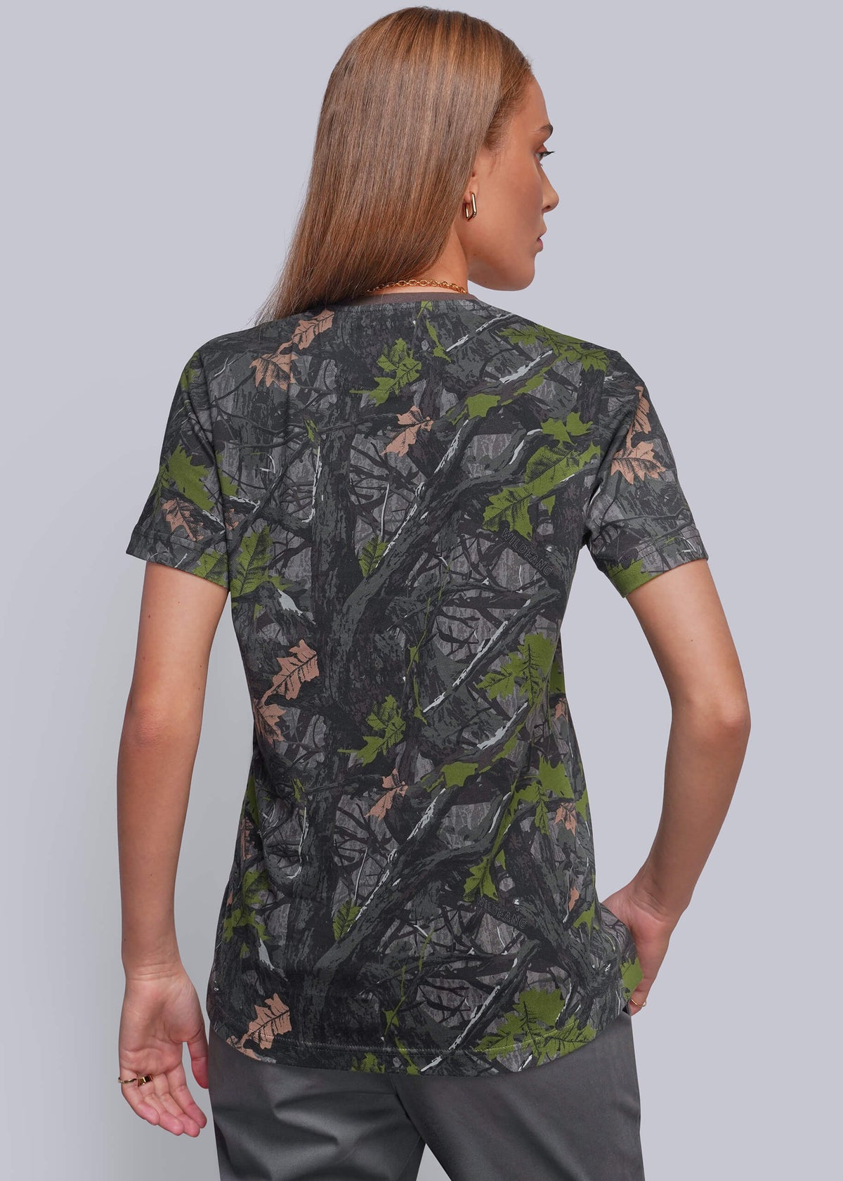 S=A woman shown from the back wearing a short-sleeve camouflage t-shirt with a realistic tree bark and leaf pattern in dark gray, green, and brown, paired with gray pants, against a solid gray studio background.