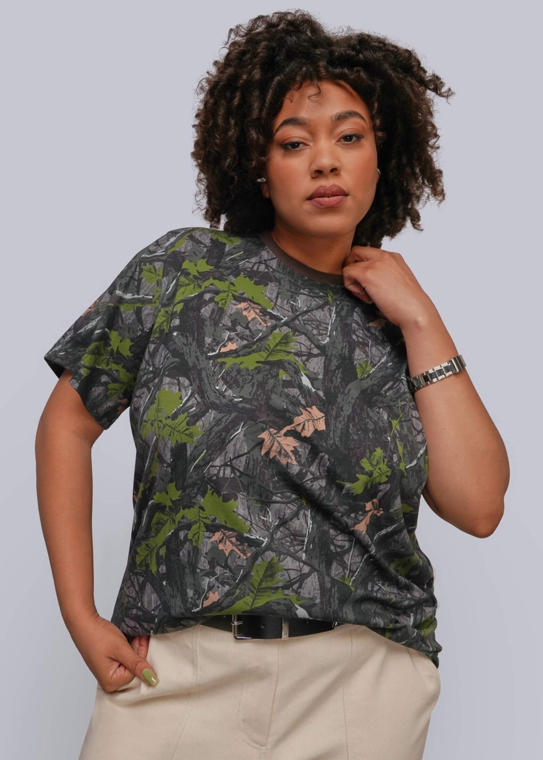 1X=A woman modeling a short-sleeve crewneck t-shirt with a dark gray and green woodland camouflage pattern, paired with beige pants, against a solid light gray background.