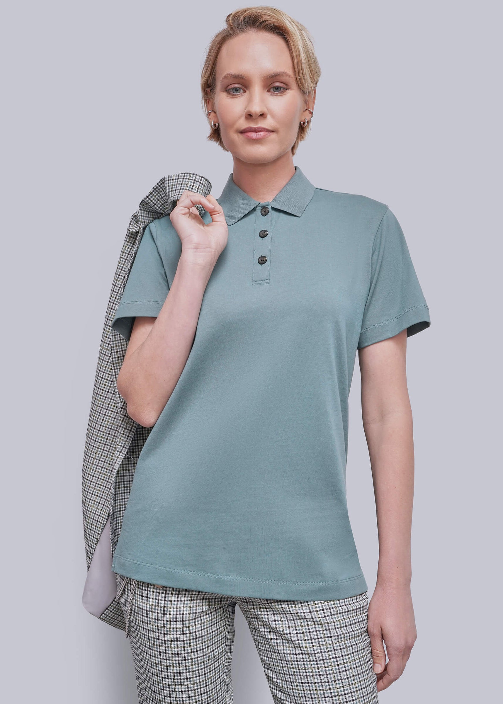 S=A woman modeling a short-sleeve polo shirt in a muted teal color, paired with matching plaid trousers and holding a plaid jacket over her shoulder, against a solid gray studio background.