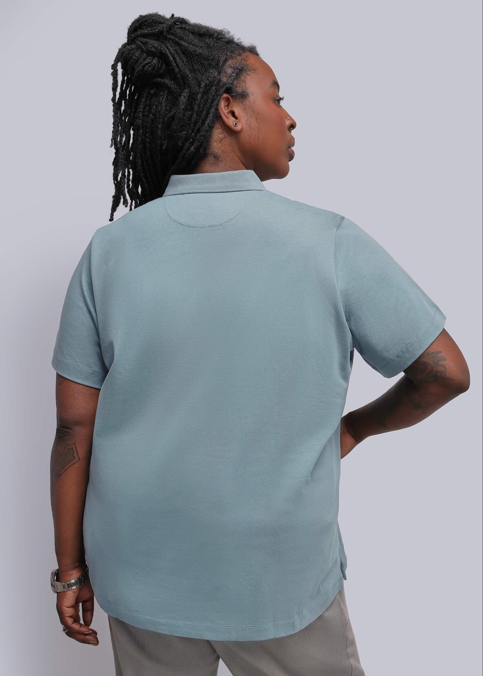 1X=A person shown from the back wearing a short-sleeve collared polo shirt in a muted light blue-gray color, paired with light beige pants, against a plain light gray studio background.